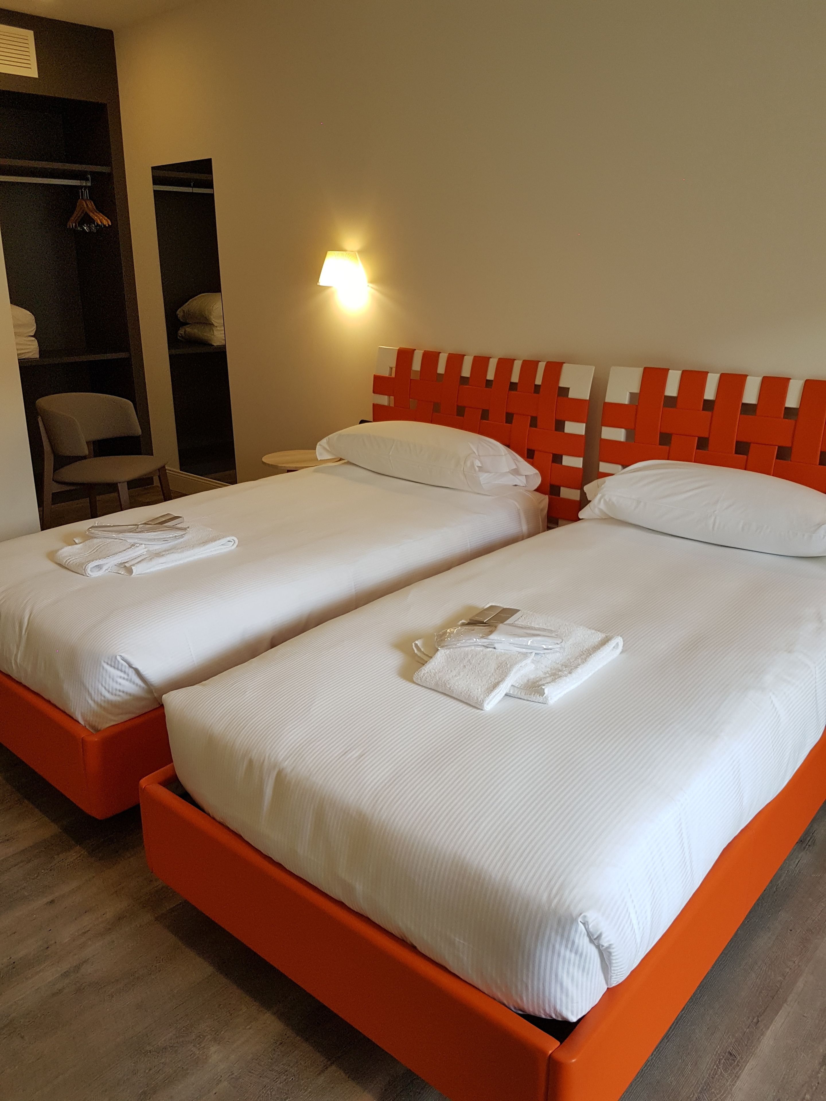 Ein Hotelzimmer mit zwei Betten, jeweils mit weißen Bettwäsche und orangefarbenen Kopfteilen. In der Ecke steht ein Sessel und ein Kleiderschrank.