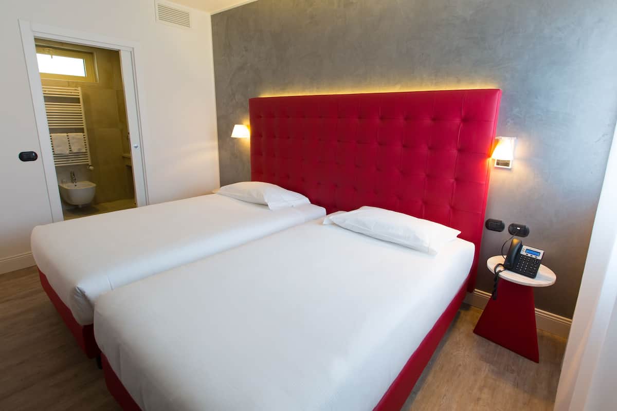 Ein Hotelzimmer mit zwei Betten, einem roten Kopfteil, Nachttischlampen, einem kleinen Tisch mit einem Telefon und einem durch eine offene Tür einsehbaren Badezimmer.