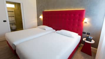 A hotel room with two beds, a red headboard, bedside lamps, a small table with a phone, and a bathroom visible through an open door.