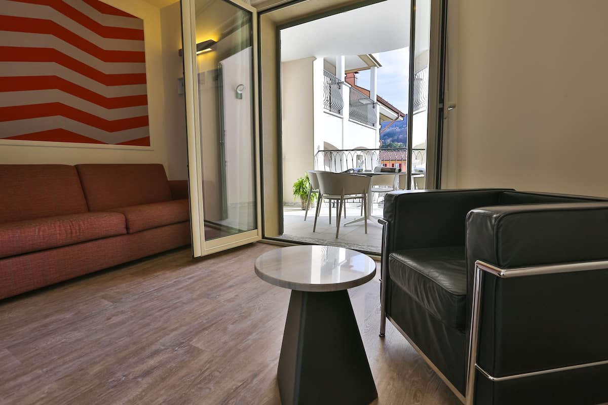 Ein modernes Wohnzimmer mit einer Couch, einem runden Tisch und einem schwarzen Ledersessel. Ein großes Fenster bietet Blick auf einen Balkon mit Gartenmöbeln.