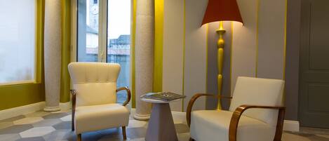 A hotel lobby with two white armchairs, a small table, a lamp, and a view of the outside through a glass door.
