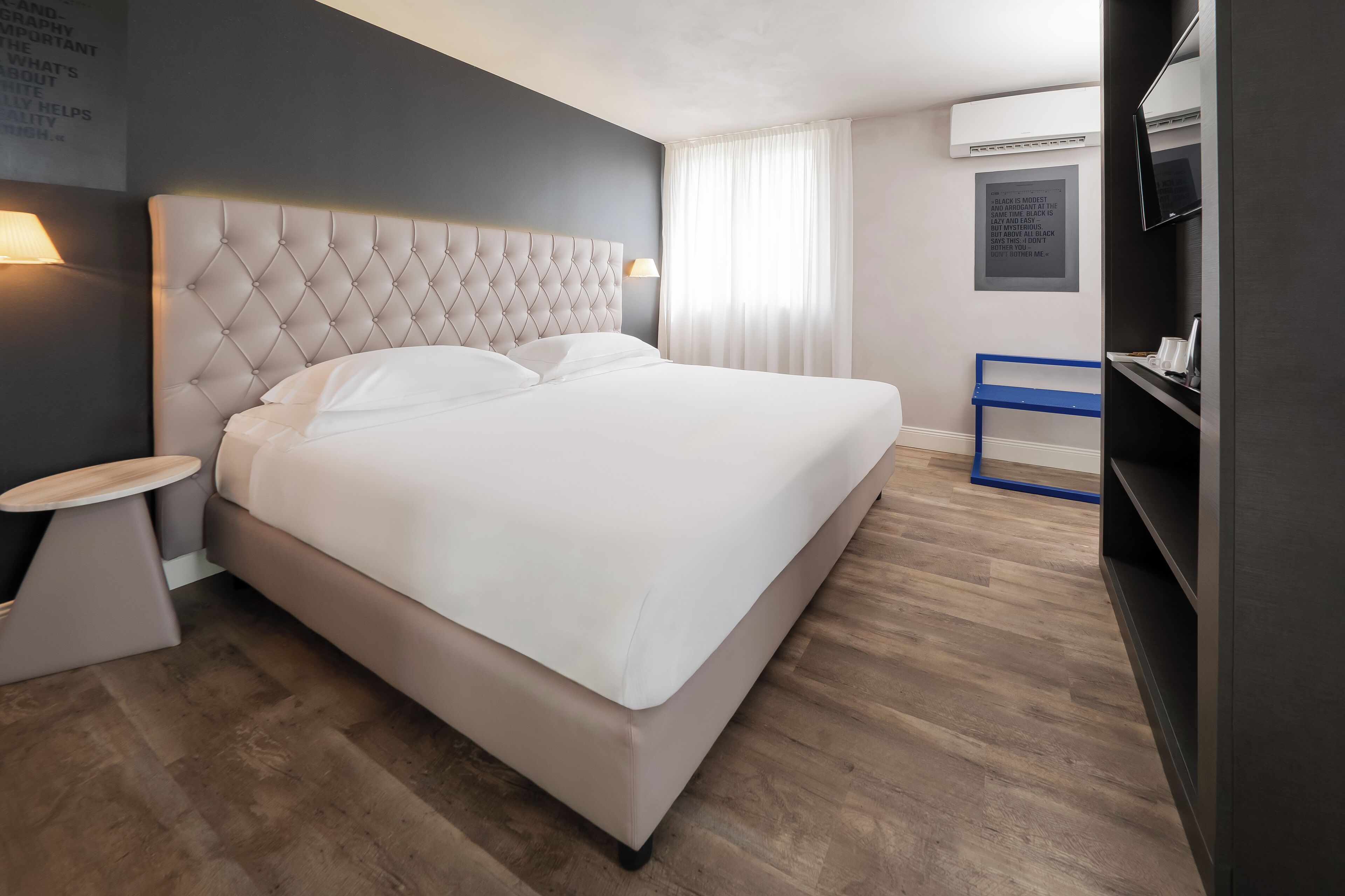 A modern hotel room with a large bed, a bedside table, a TV, and a wall-mounted air conditioner.
