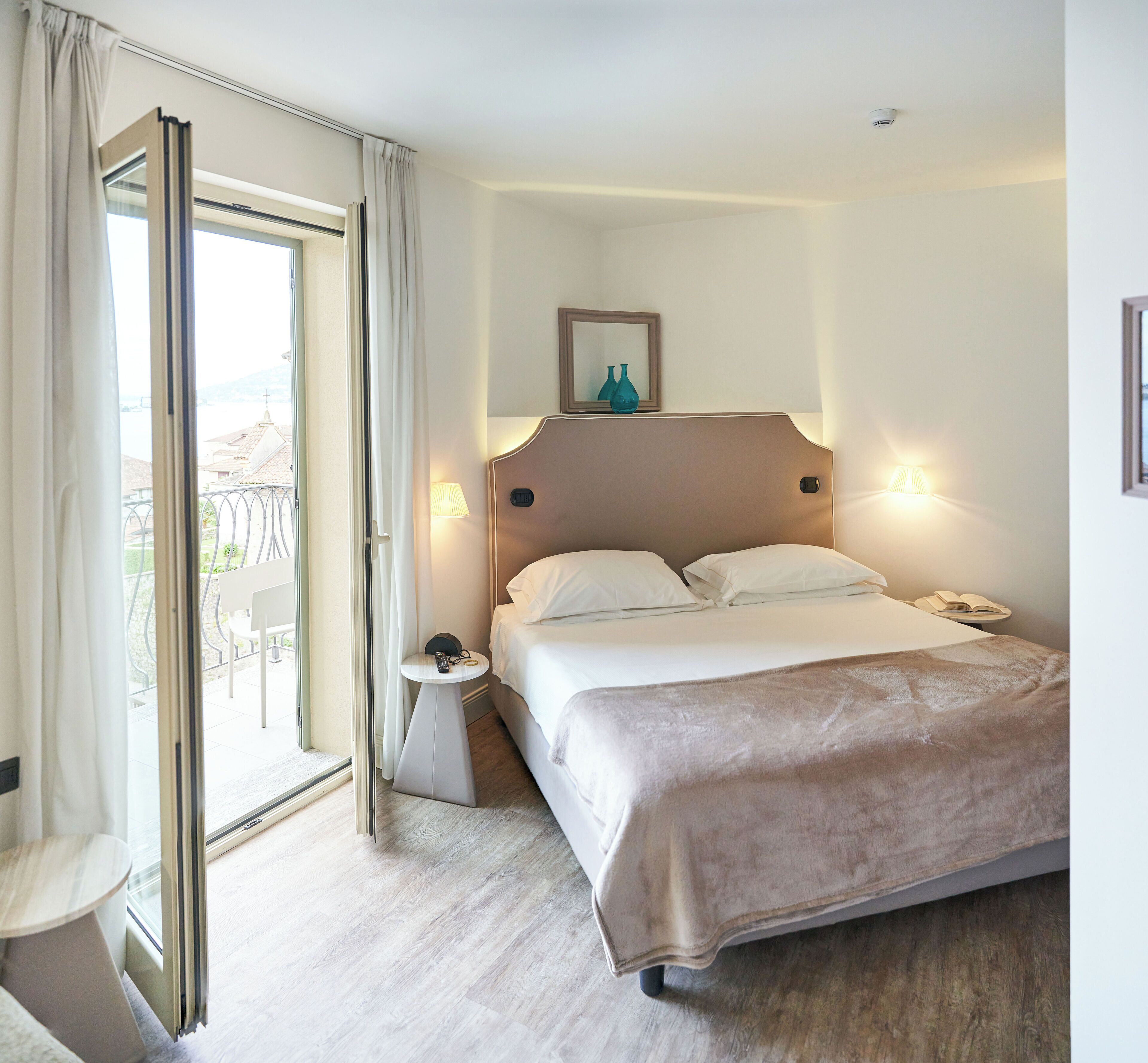 A hotel room with a bed, bedside lamps, a small table with a remote control, and a view through a large window.