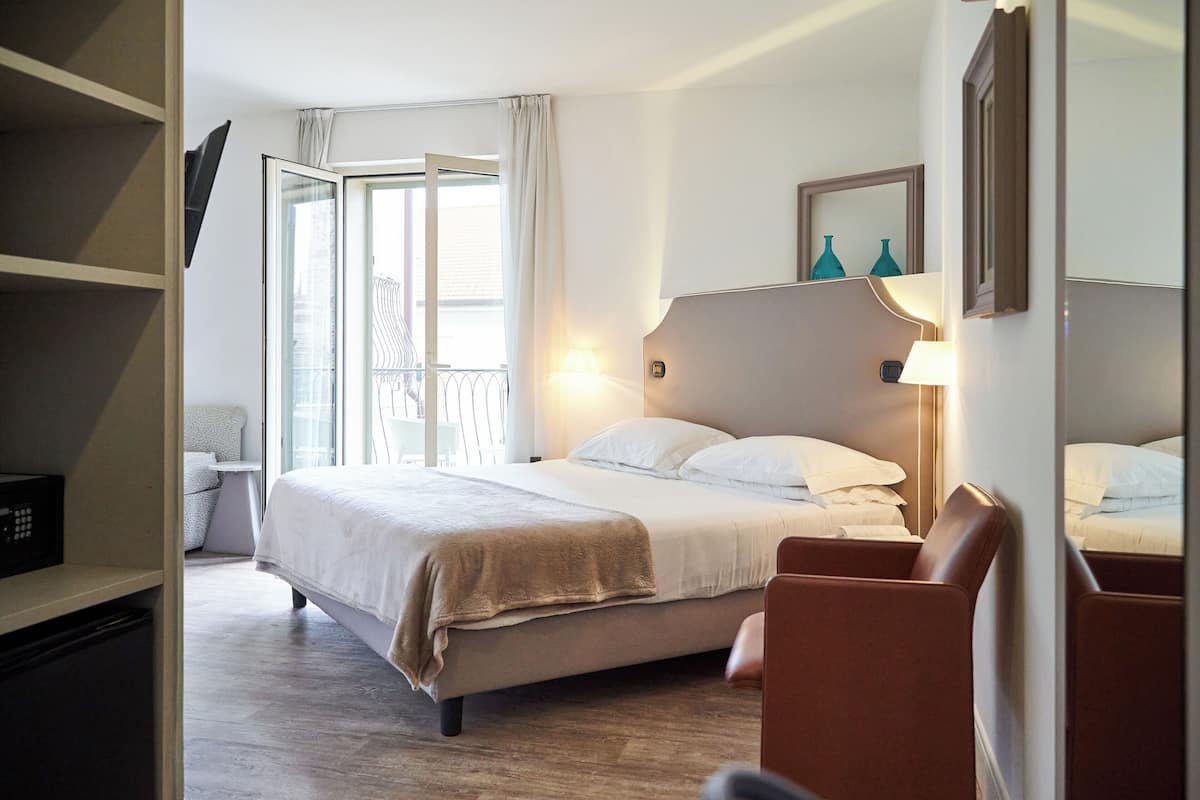 Ein Hotelzimmer mit Bett, Sessel, Fernseher und einem Balkon mit Geländer.