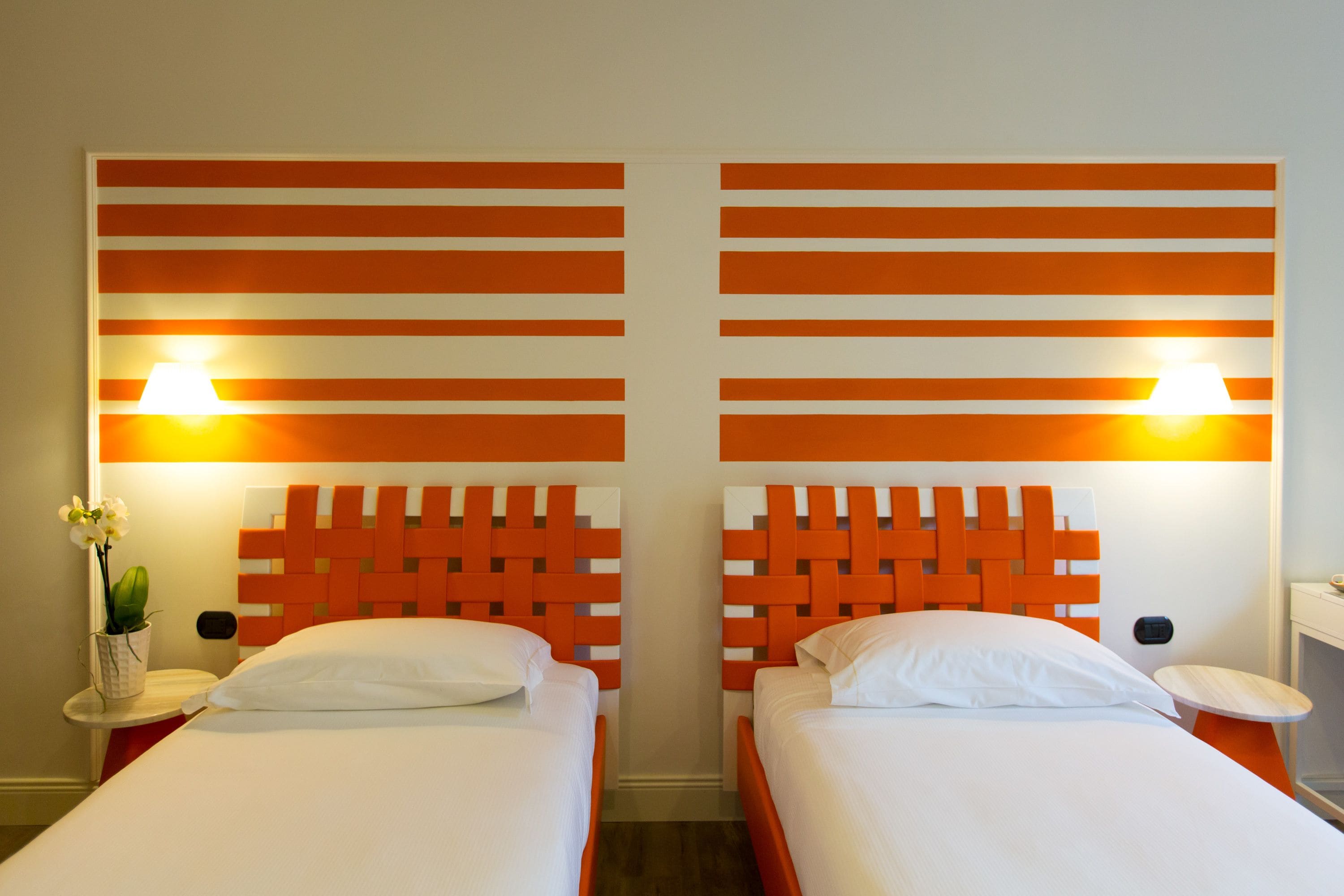 A hotel room with two beds, each with a headboard featuring a geometric pattern. There are bedside tables with lamps and a small vase with flowers on one side.