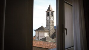 Vista pela janela de um edifício com torre de relógio, cruz no topo e telhado de azulejos.