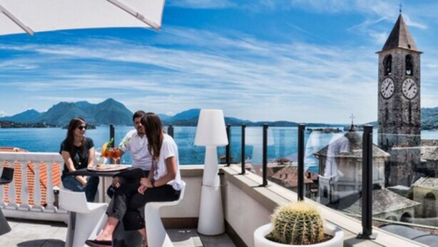 Terraço com mesa e cadeiras, um vaso de planta e vista para um corpo de água e montanhas ao fundo.