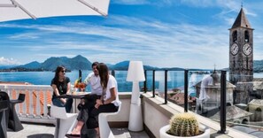 Una terraza con mesa y sillas, una planta en maceta y vistas a un cuerpo de agua y montañas al fondo.