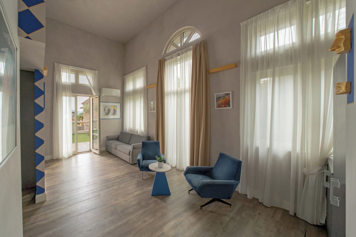 Ein modernes Wohnzimmer mit einer Couch, zwei Sesseln, einem Couchtisch und großen Fenstern mit leicht transparenten Vorhängen.