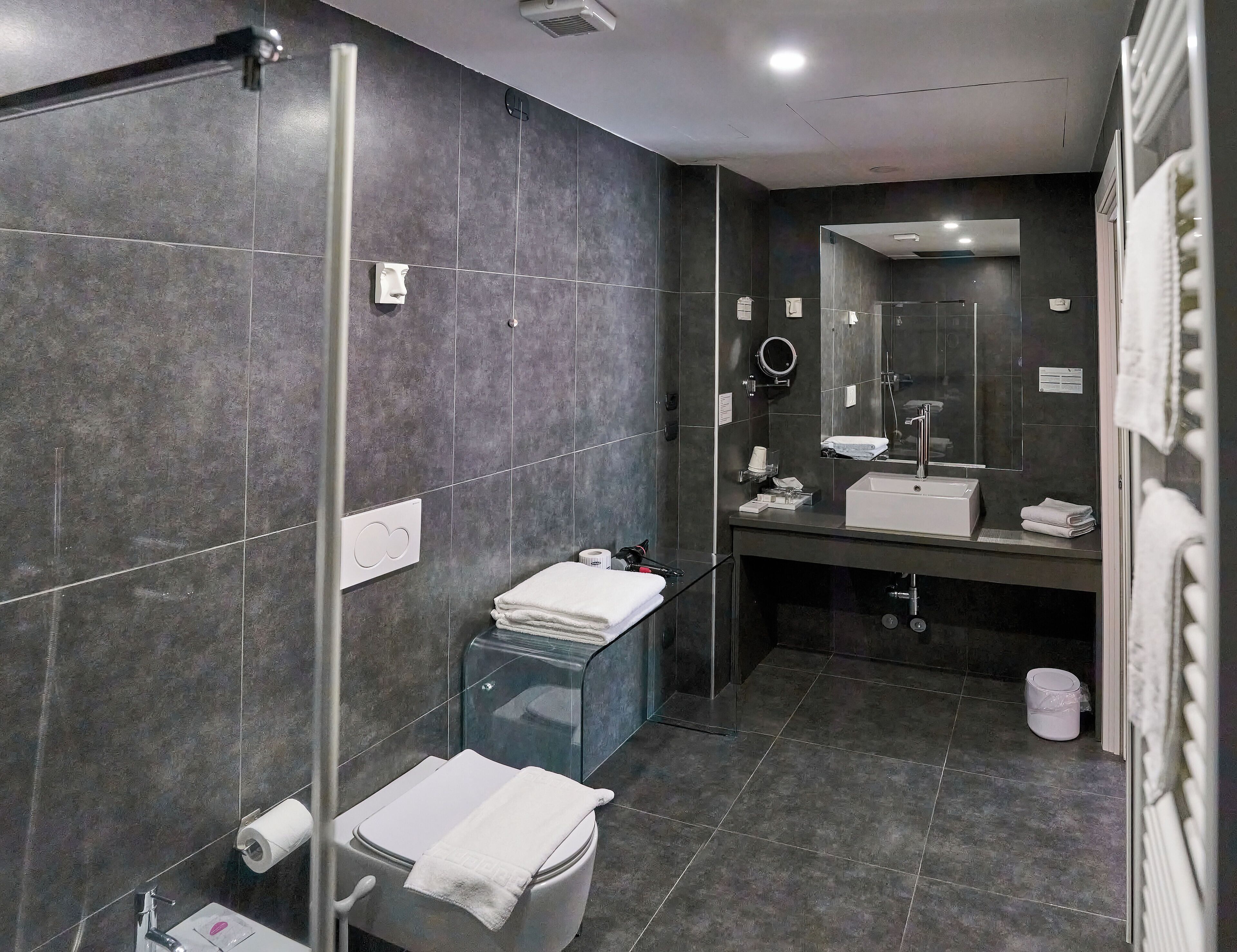 Un bagno moderno con piastrelle scure, cabina doccia in vetro, lavabo bianco e uno specchio grande.