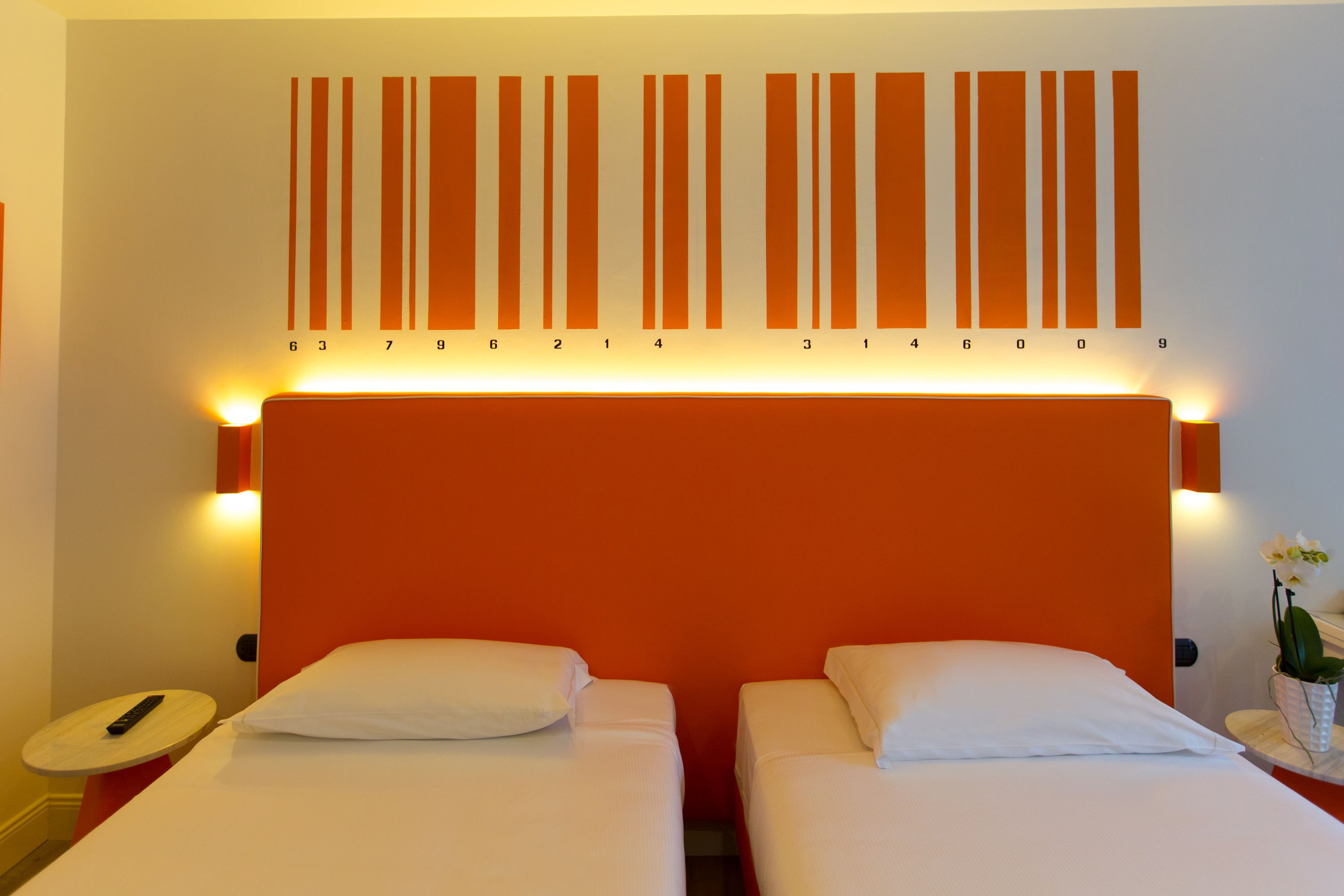 Ein Hotelzimmer mit zwei Betten, einem großen orangefarbenen Kopfteil und einer Wand mit vertikalen, orangefarbenen Streifen und Nummern.