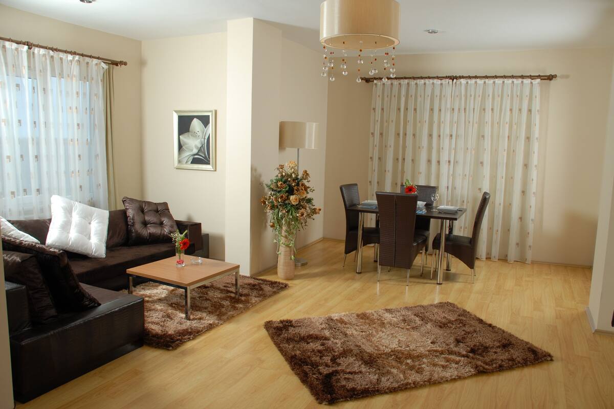 A living room with a brown sofa, a coffee table, a dining table with chairs, a lamp, and a large window with curtains.