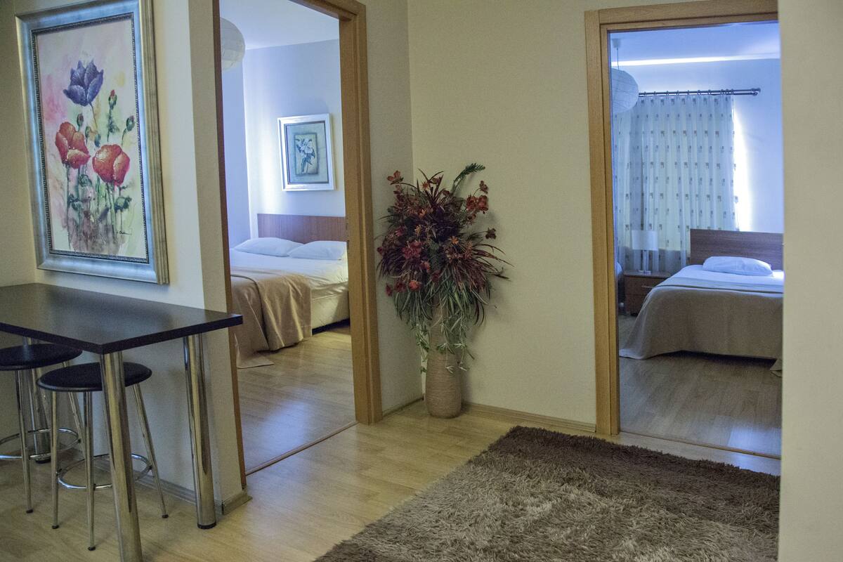 A hotel room with a bed, a desk with chairs, a large plant, and a painting on the wall.