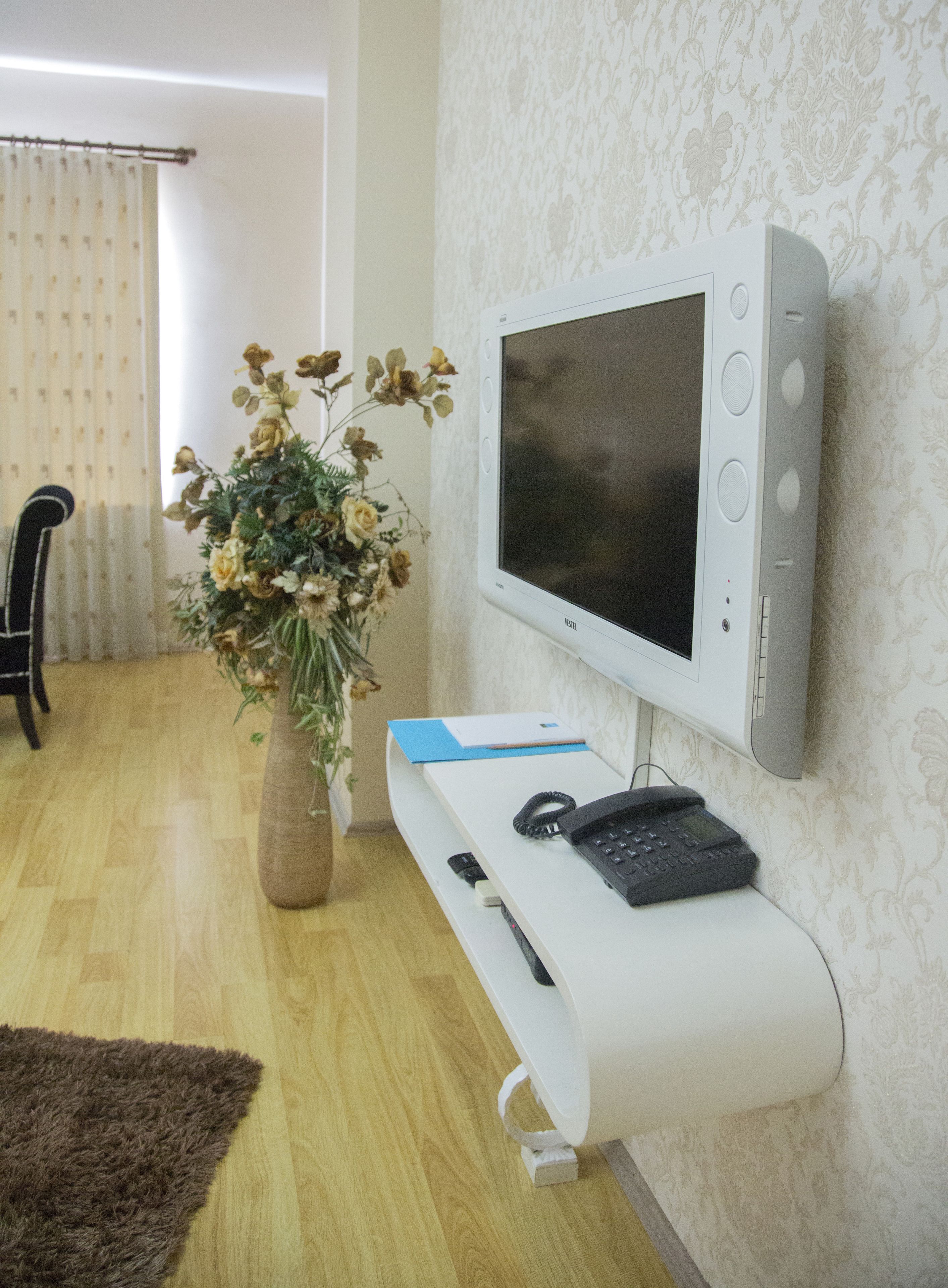 Een moderne woonkamer met een flatscreen televisie aan de muur, een witte console tafel met een telefoon en afstandsbediening, een vaas met gedroogde bloemen en een houten vloer.