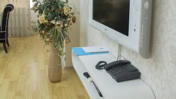 Een moderne woonkamer met een flatscreen televisie aan de muur, een witte console tafel met een telefoon en afstandsbediening, een vaas met gedroogde bloemen en een houten vloer.