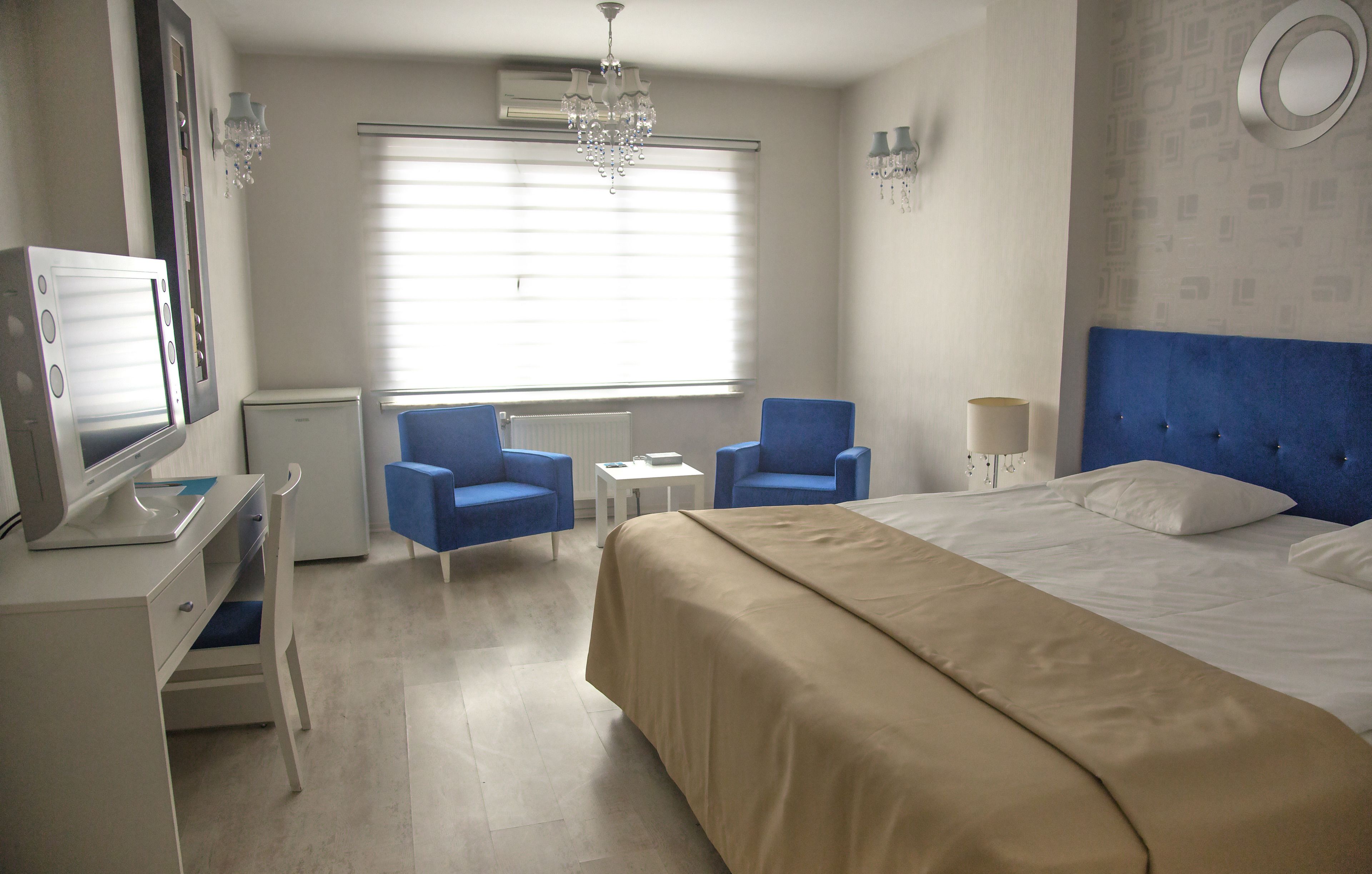 Una habitación de hotel con una cama, dos sillones azules, un escritorio con una computadora y un candelabro.