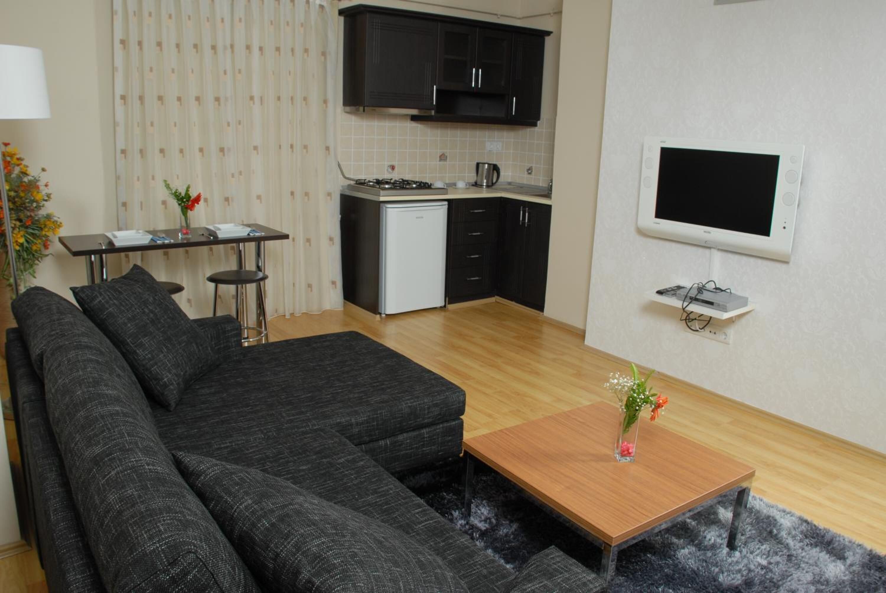 A modern living room with a sectional sofa, a wooden coffee table, and a small kitchen area with a refrigerator, stove, and microwave.