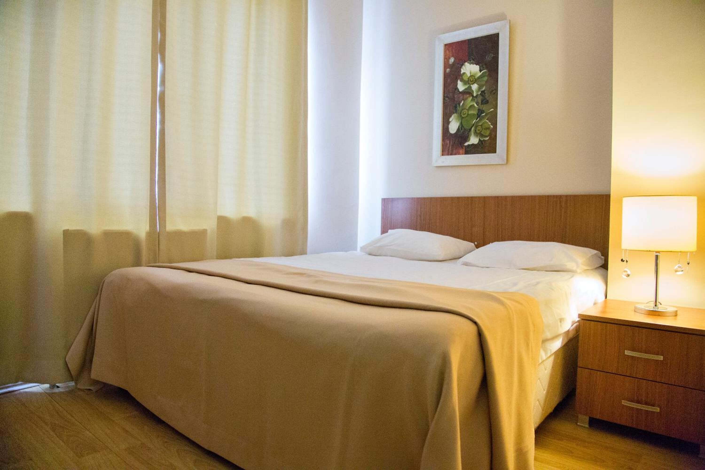 Habitación de hotel con cama, mesita de noche con lámpara, un cuadro en la pared y ventana con cortinas.