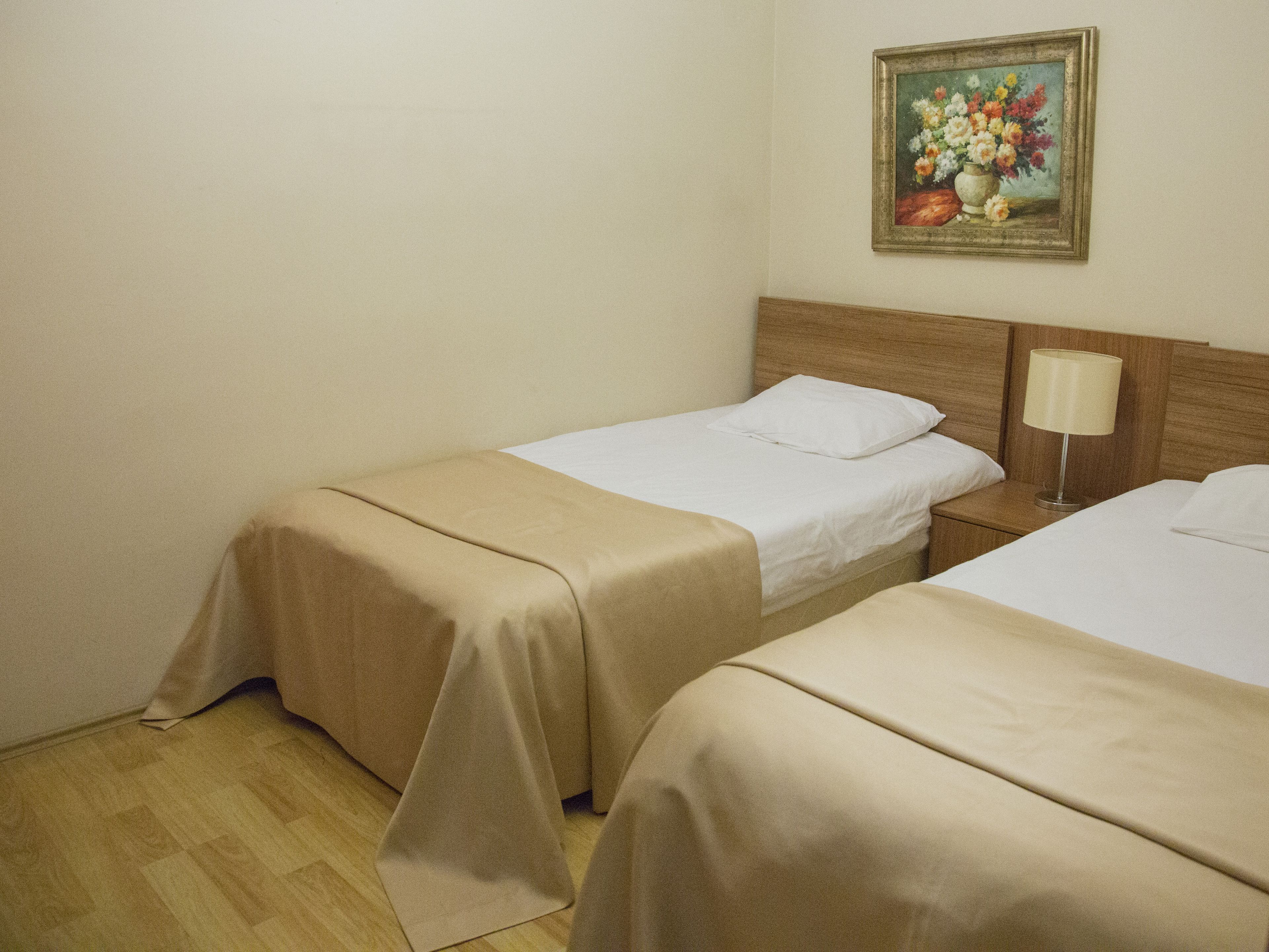 A hotel room with two single beds, a wooden headboard, a nightstand with a lamp, and a painting of flowers on the wall.