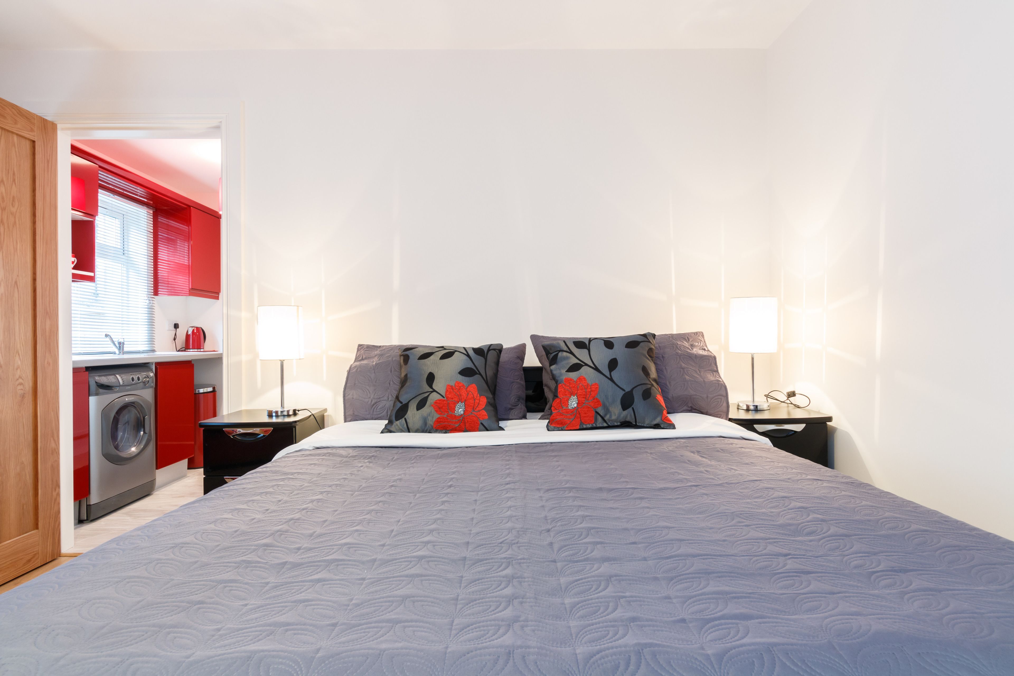 A bedroom with a bed, two nightstands, a lamp, a red washing machine, and a red and white kitchen area.