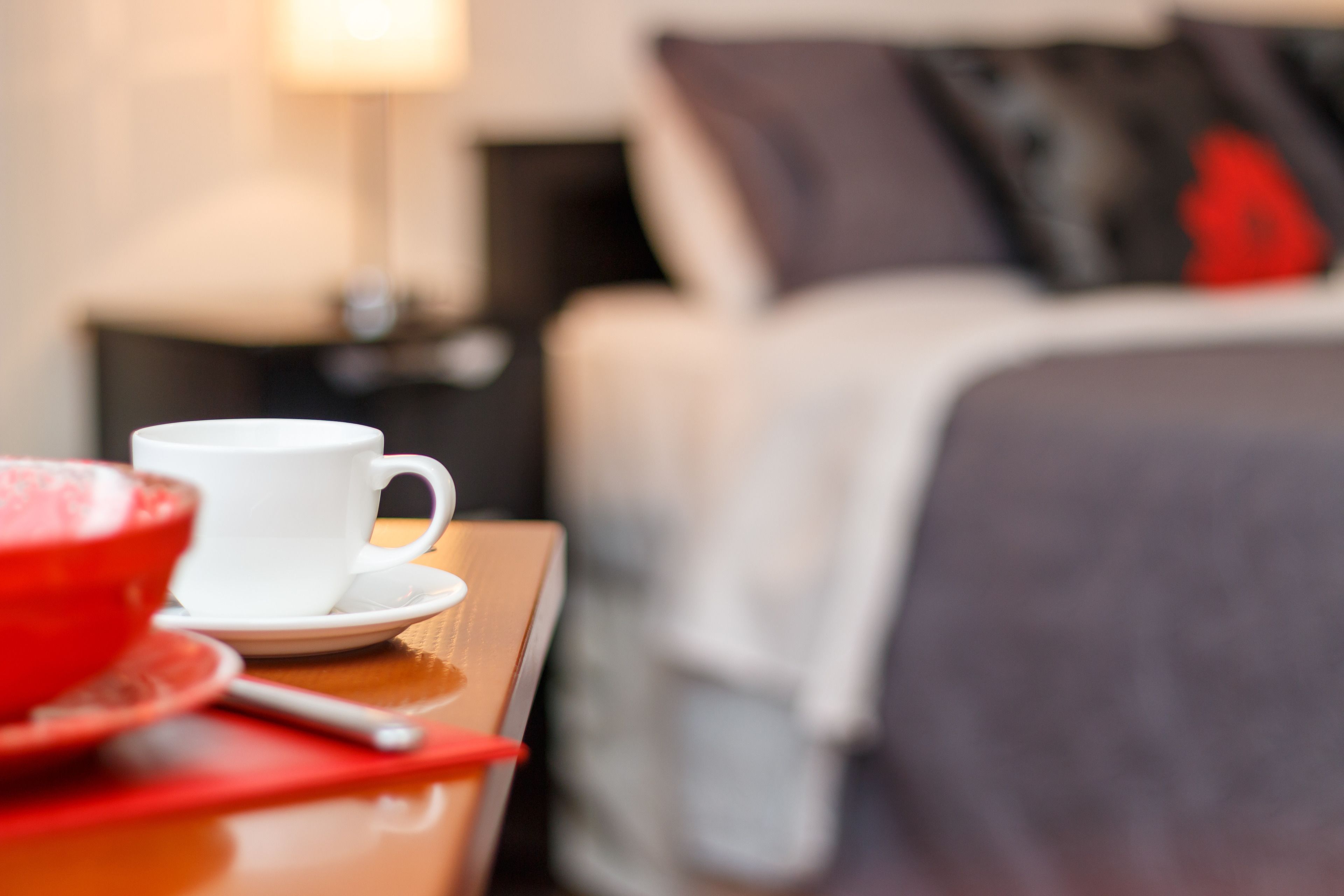 A hotel room with a bed, a bedside table with a white cup and saucer, a red bowl, and a spoon.