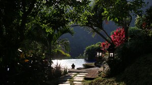 A stone pathway leading to a small wooden structure with a lantern, surrounded by lush greenery and a body of water in the background. - Villa Patria (Sukasada)