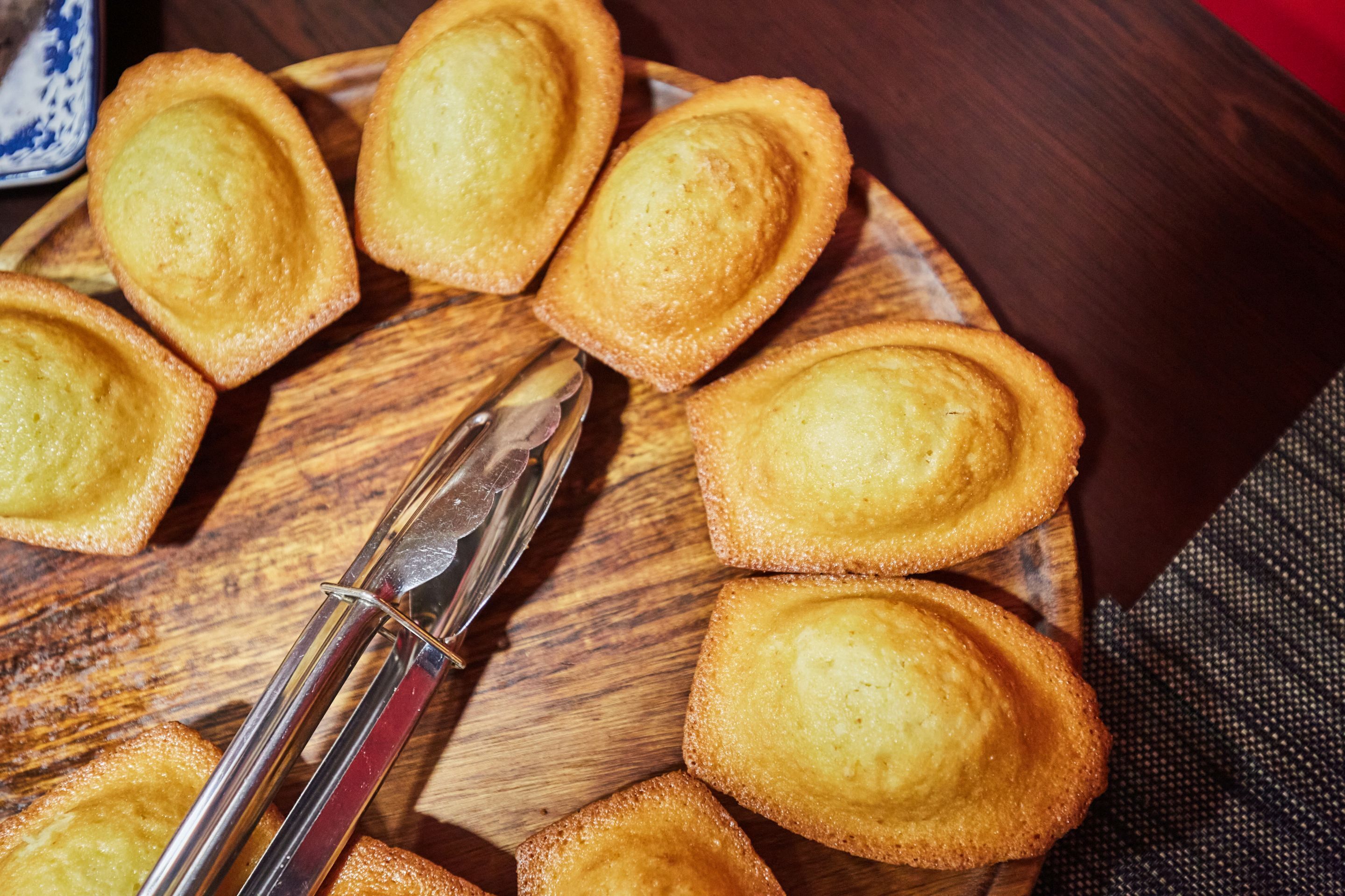 A wooden plate with several golden-brown pastries, a knife, and a pair of tongs on a table.