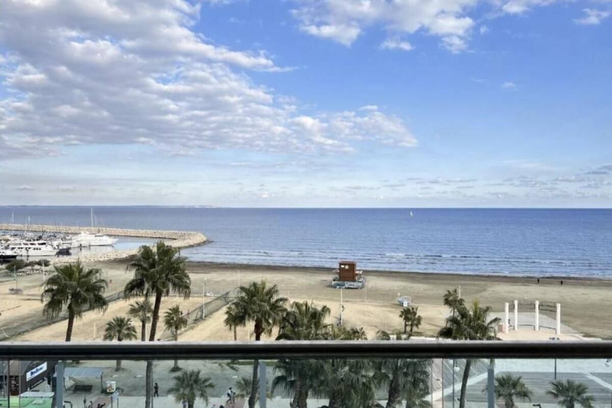 Vista a la playa desde un balcón, con palmeras, arena y un muelle con barcos.