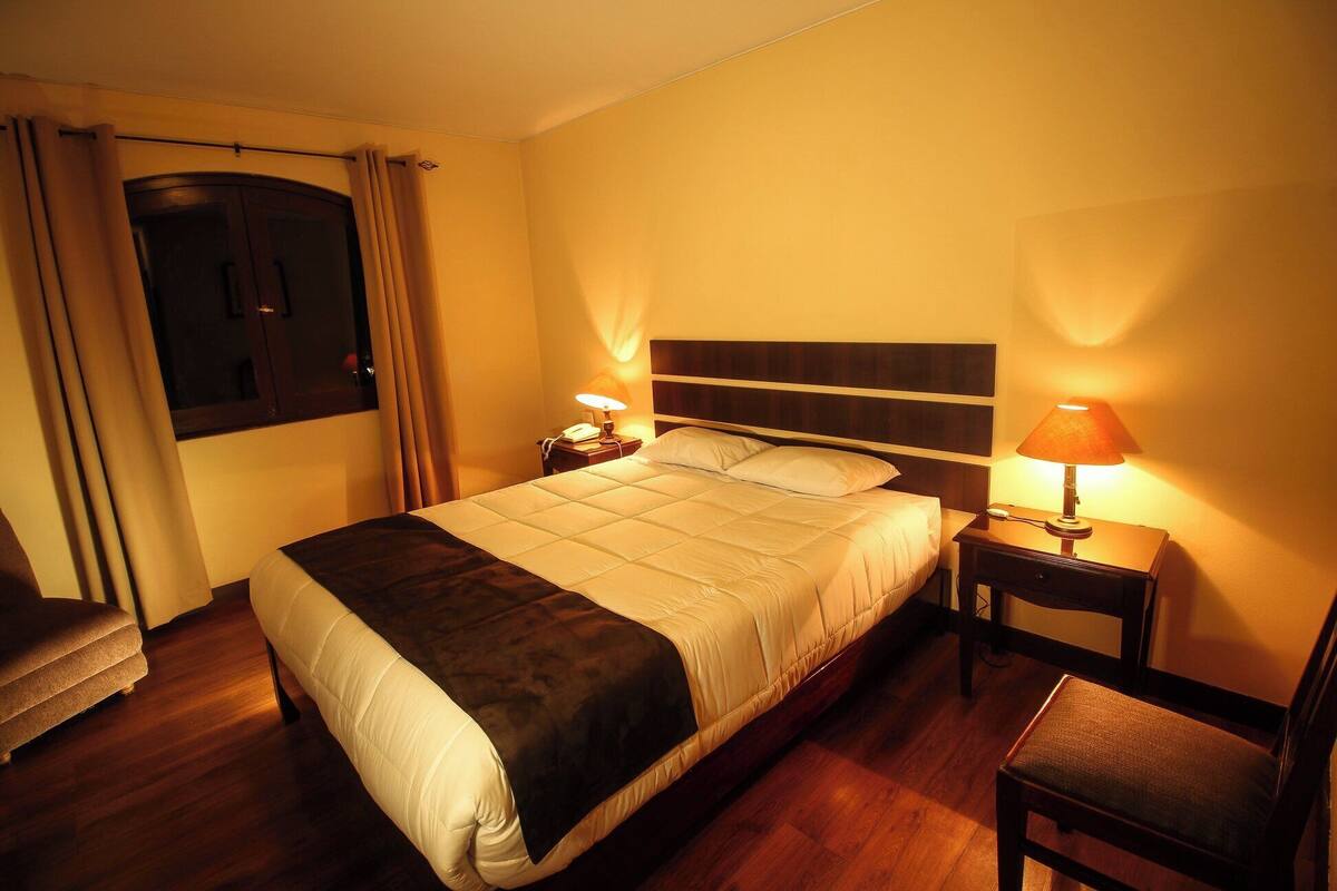 Una habitación de hotel con cama, lámparas de noche, una silla y un ventanal con cortinas.