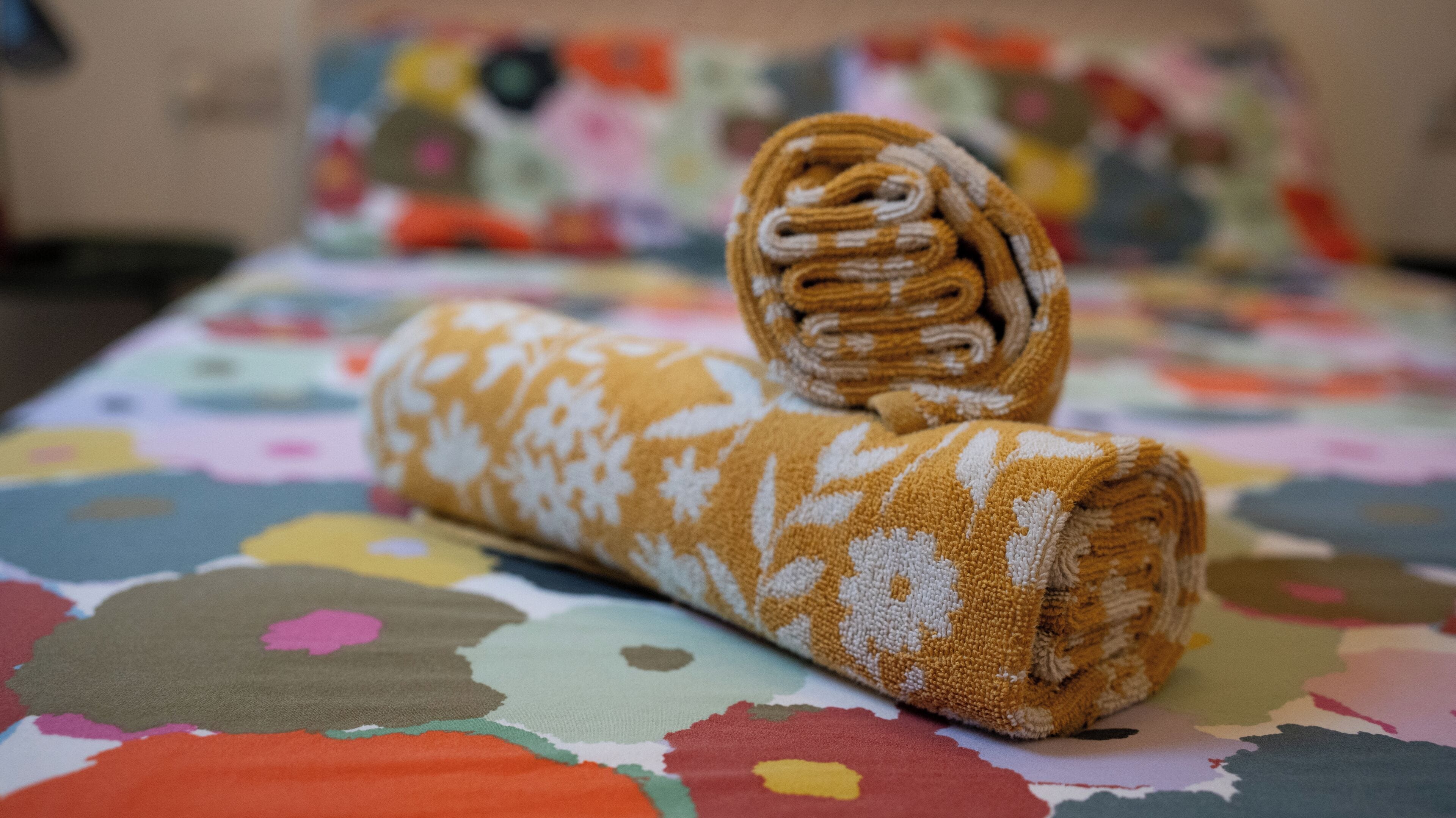 A rolled towel on a bed with a multicolored floral bedspread.