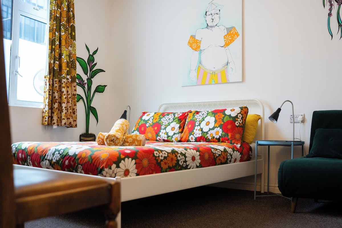 A bed with floral bedding, a bedside table, a lamp, a green armchair, and a potted plant.