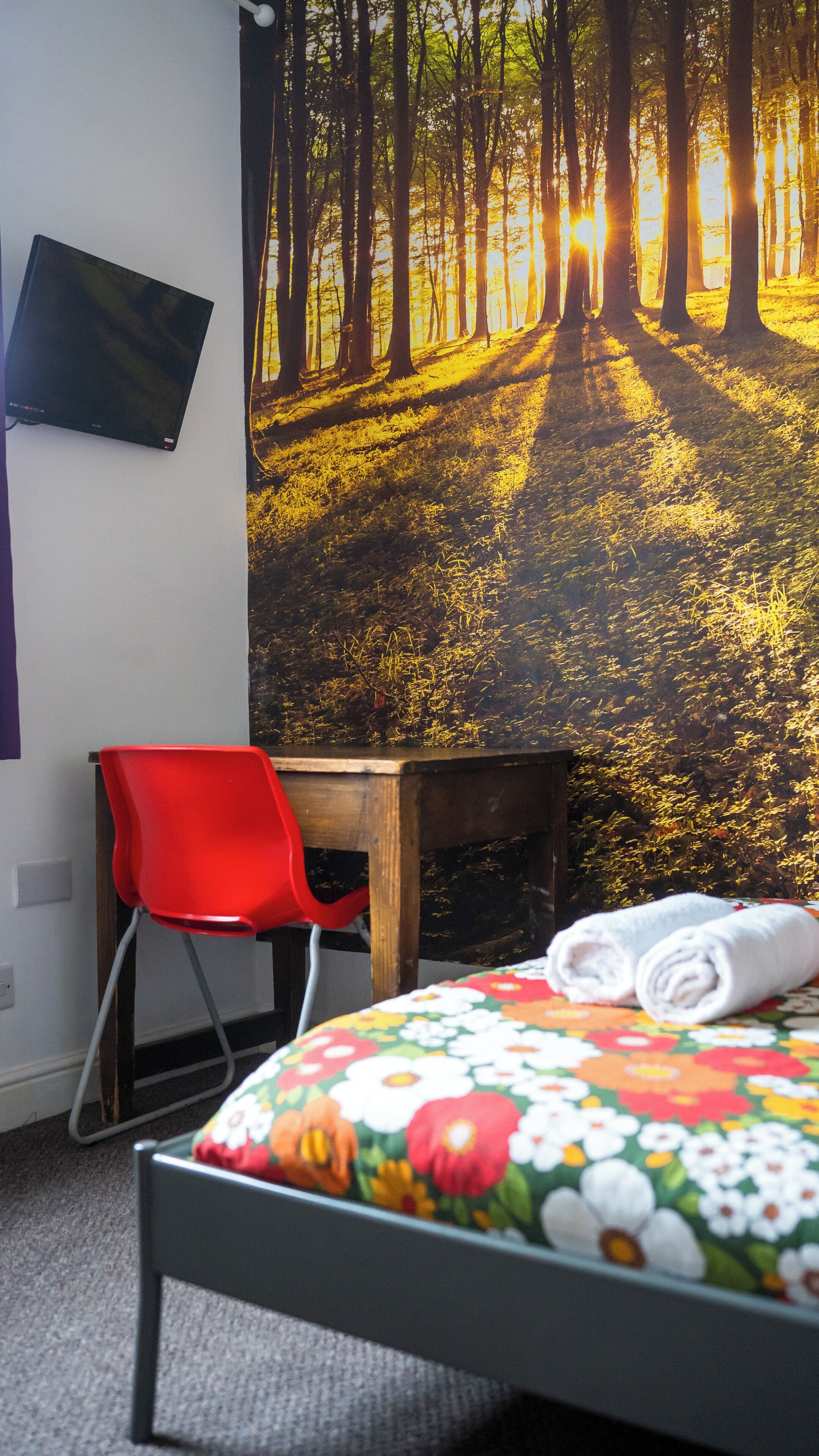 A bedroom with a bed, a red chair, a wooden table, a TV on the wall, and a forest scene wallpaper.