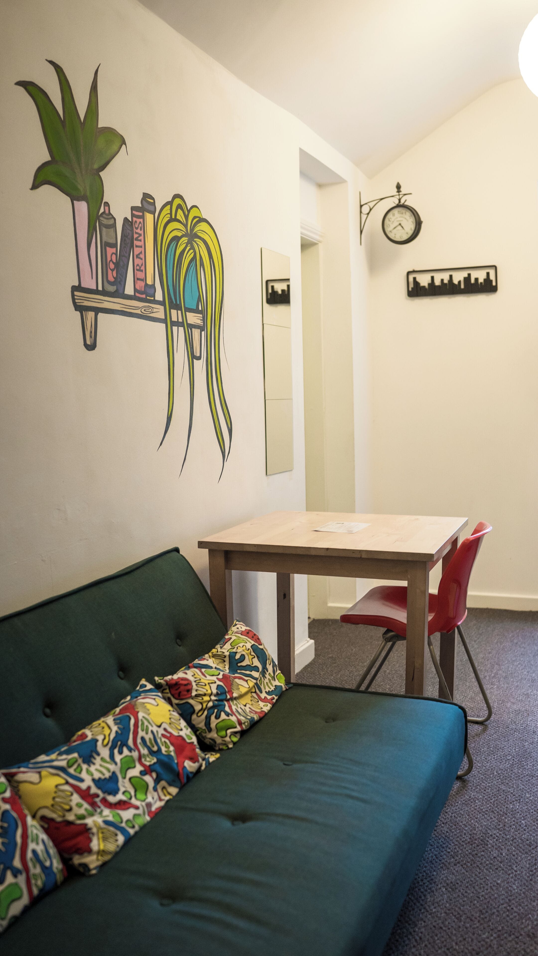 Una habitación con un sofá verde, una mesa de madera, una silla roja, un reloj de pared y una decoración con el perfil de un horizonte urbano.