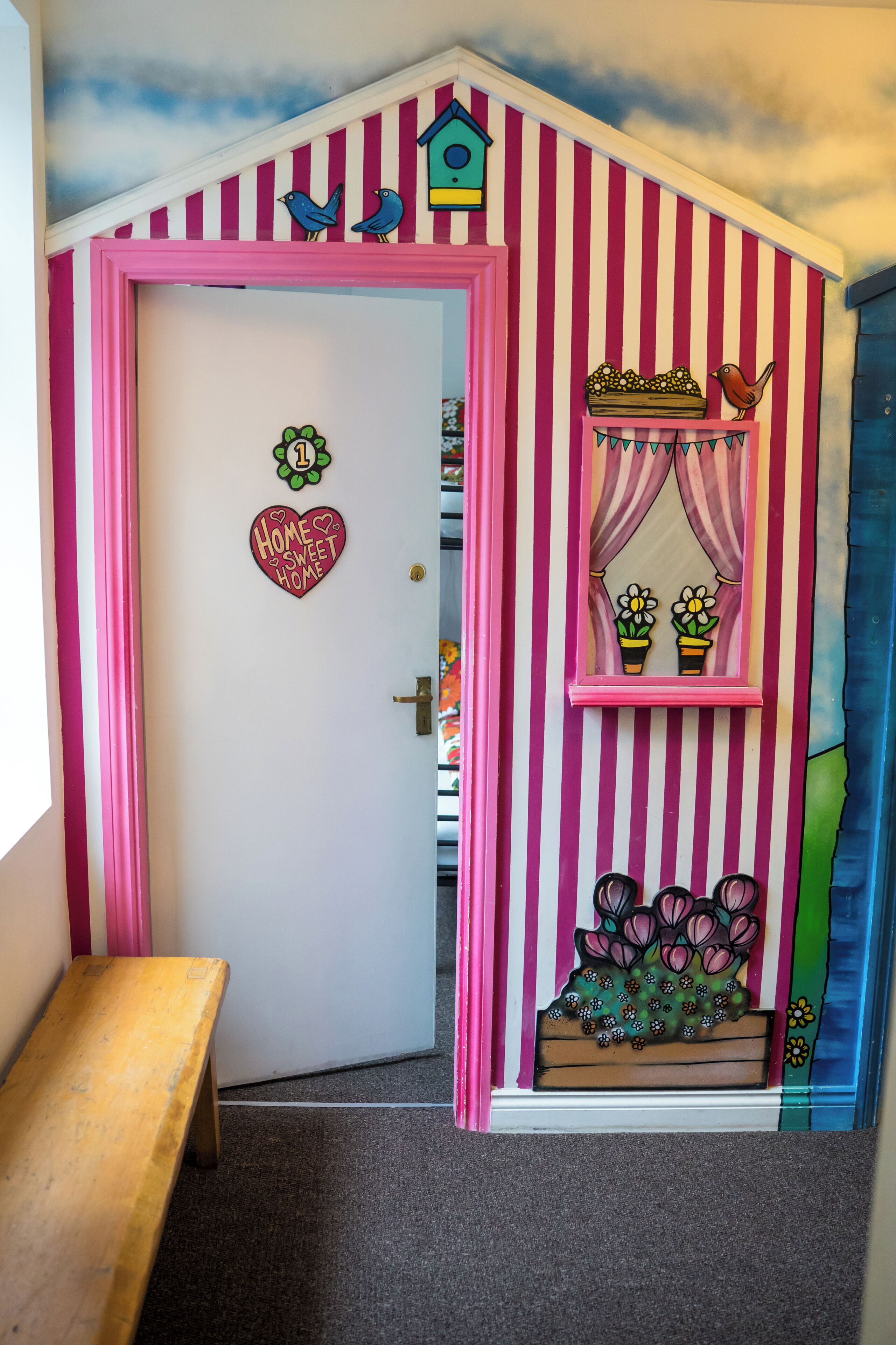 Una habitación con una pared a rayas, una puerta con un letrero con forma de corazón, un banco y una ventana con cortinas.
