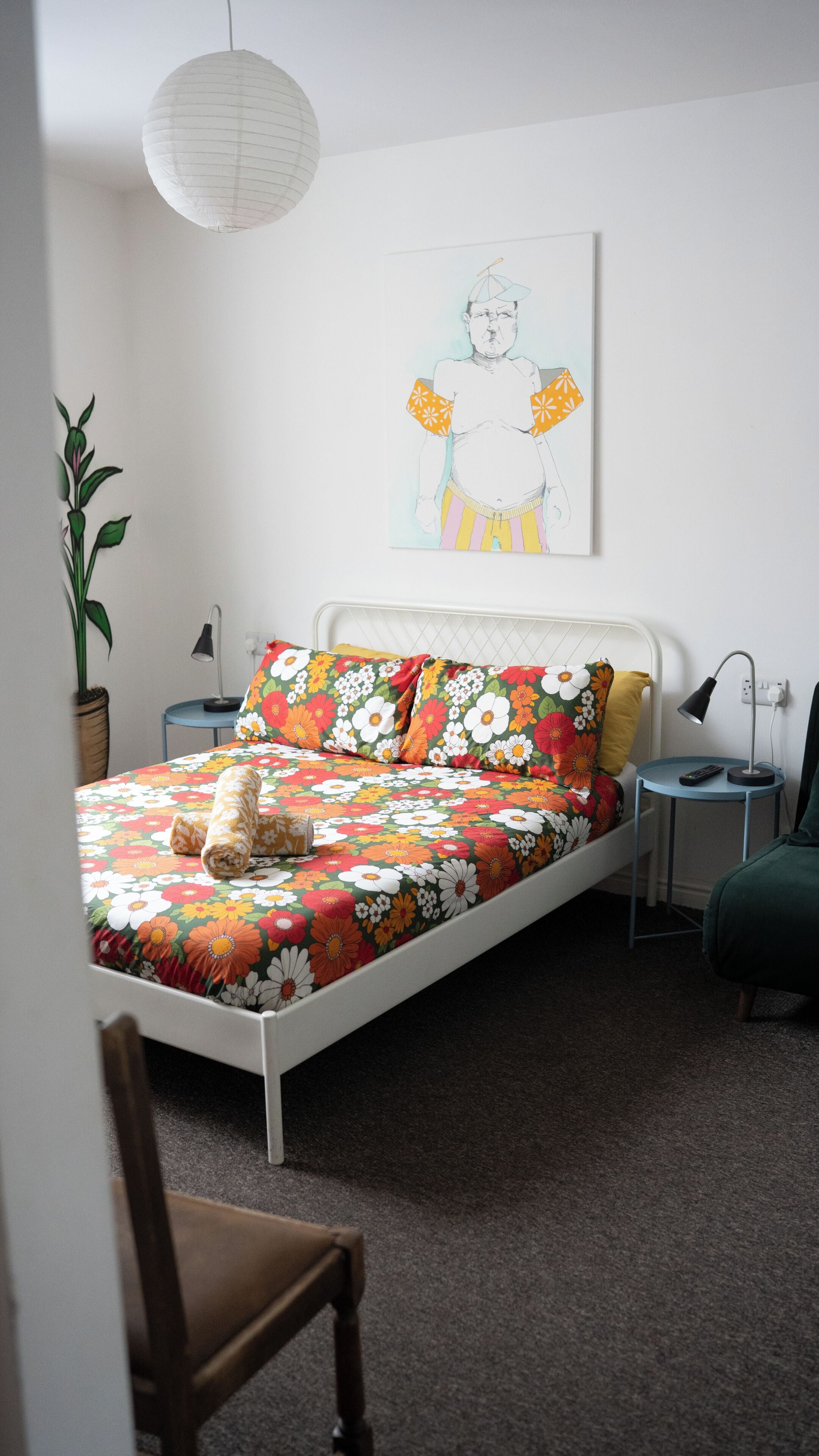 A bed with floral bedding, a bedside lamp, a nightstand, a potted plant, and a wall-mounted artwork.