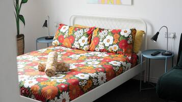 A bed with floral bedding, a bedside lamp, a nightstand, a potted plant, and a wall-mounted artwork.