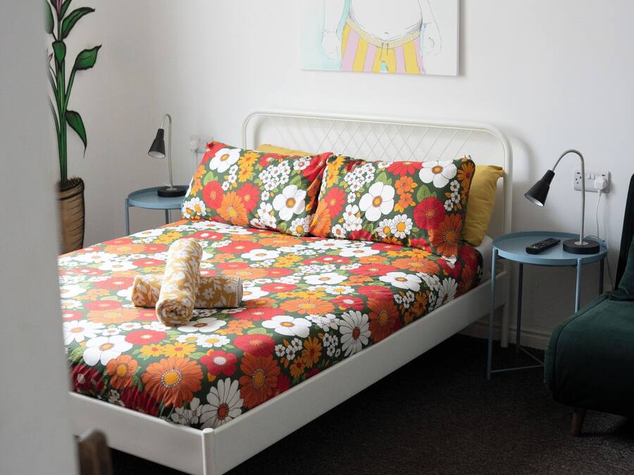 A bed with floral bedding, a bedside lamp, a nightstand, a potted plant, and a wall-mounted artwork.
