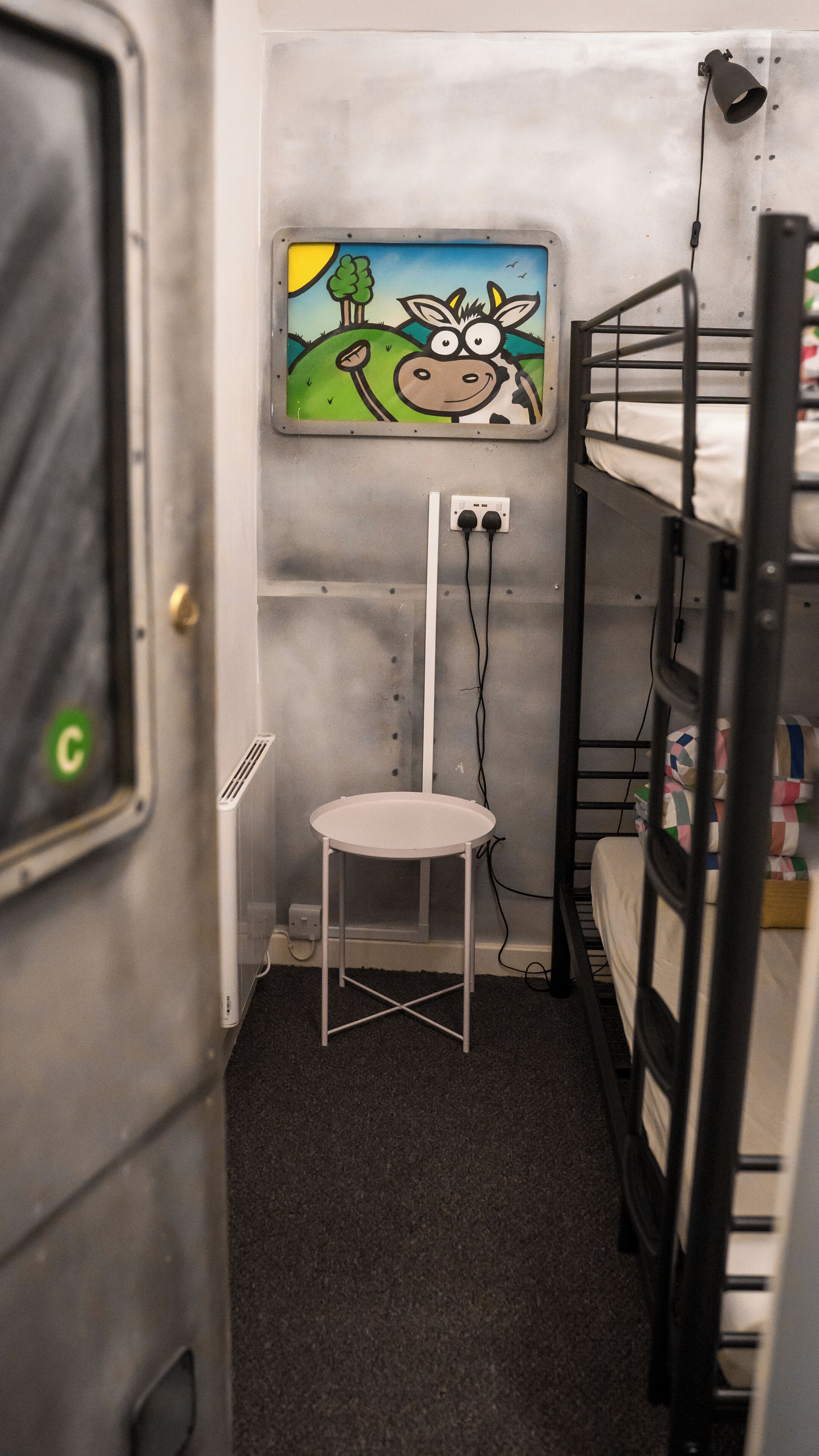 Una habitación pequeña y estrecha con literas, una mesa blanca y un enchufe en la pared.