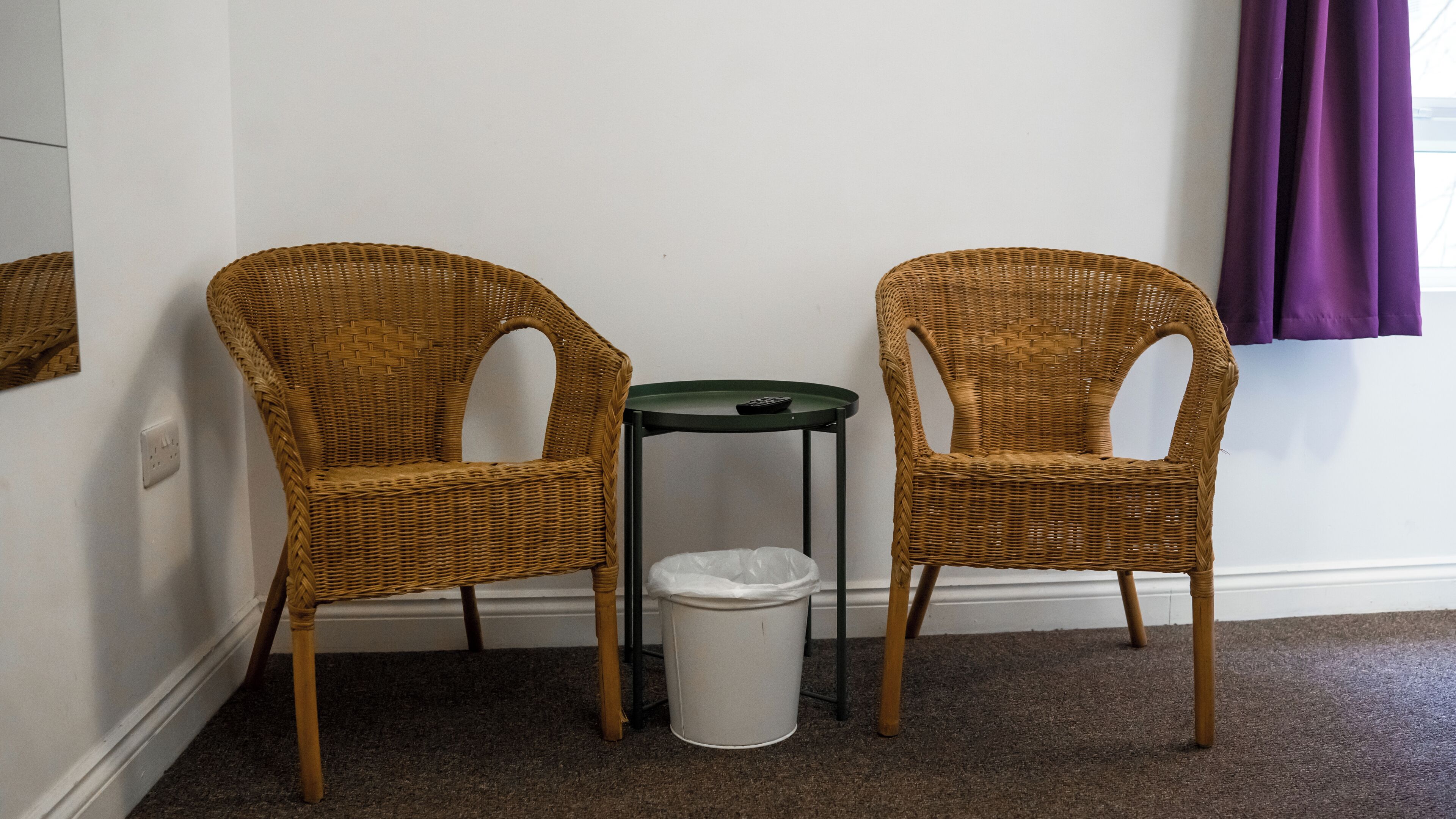 Two wicker chairs, a small trash bin, and a green table against a white wall with purple curtains.