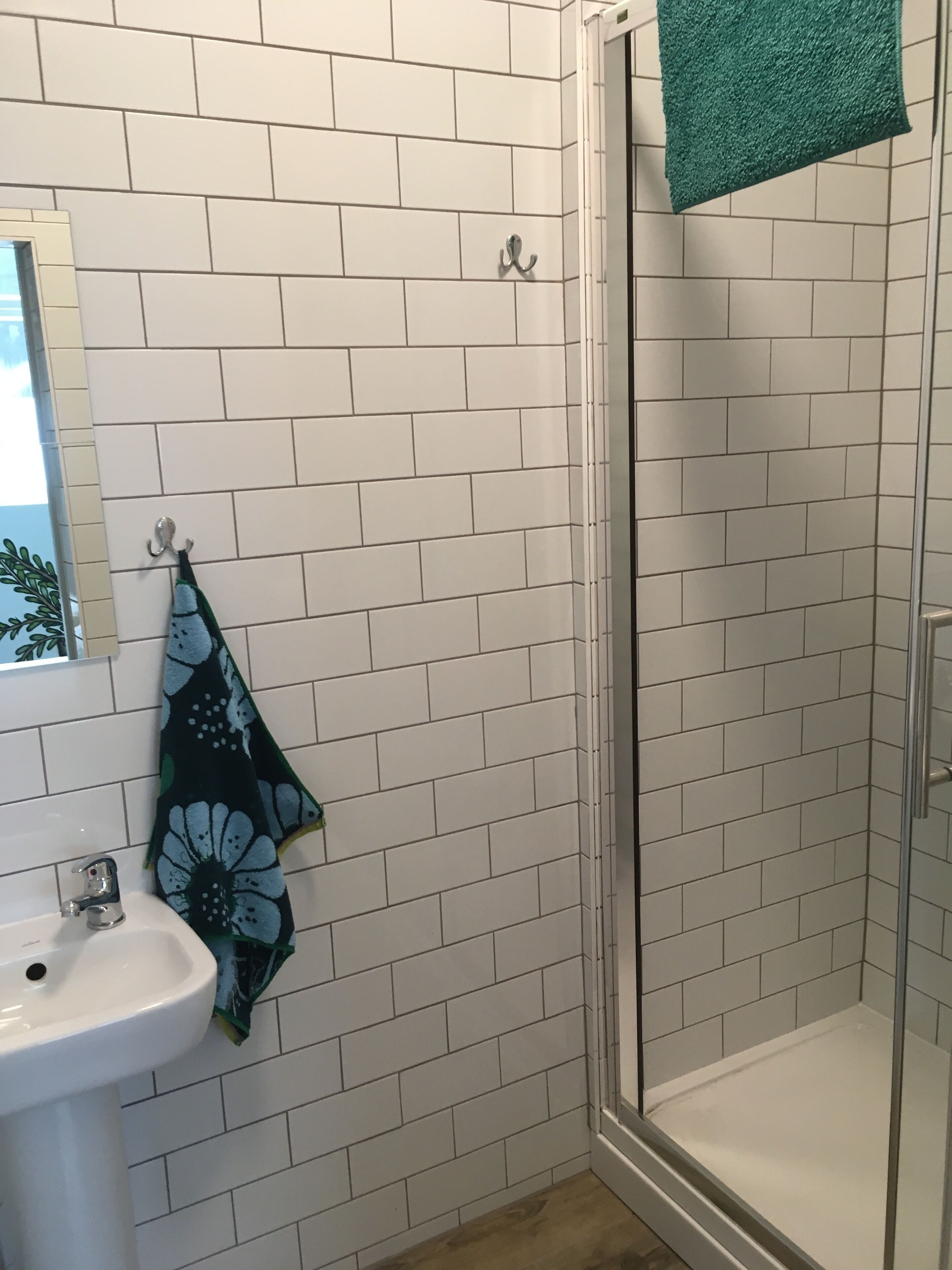 A bathroom with white tiled walls, a glass shower enclosure, and a sink with a green floral towel hanging on the wall.