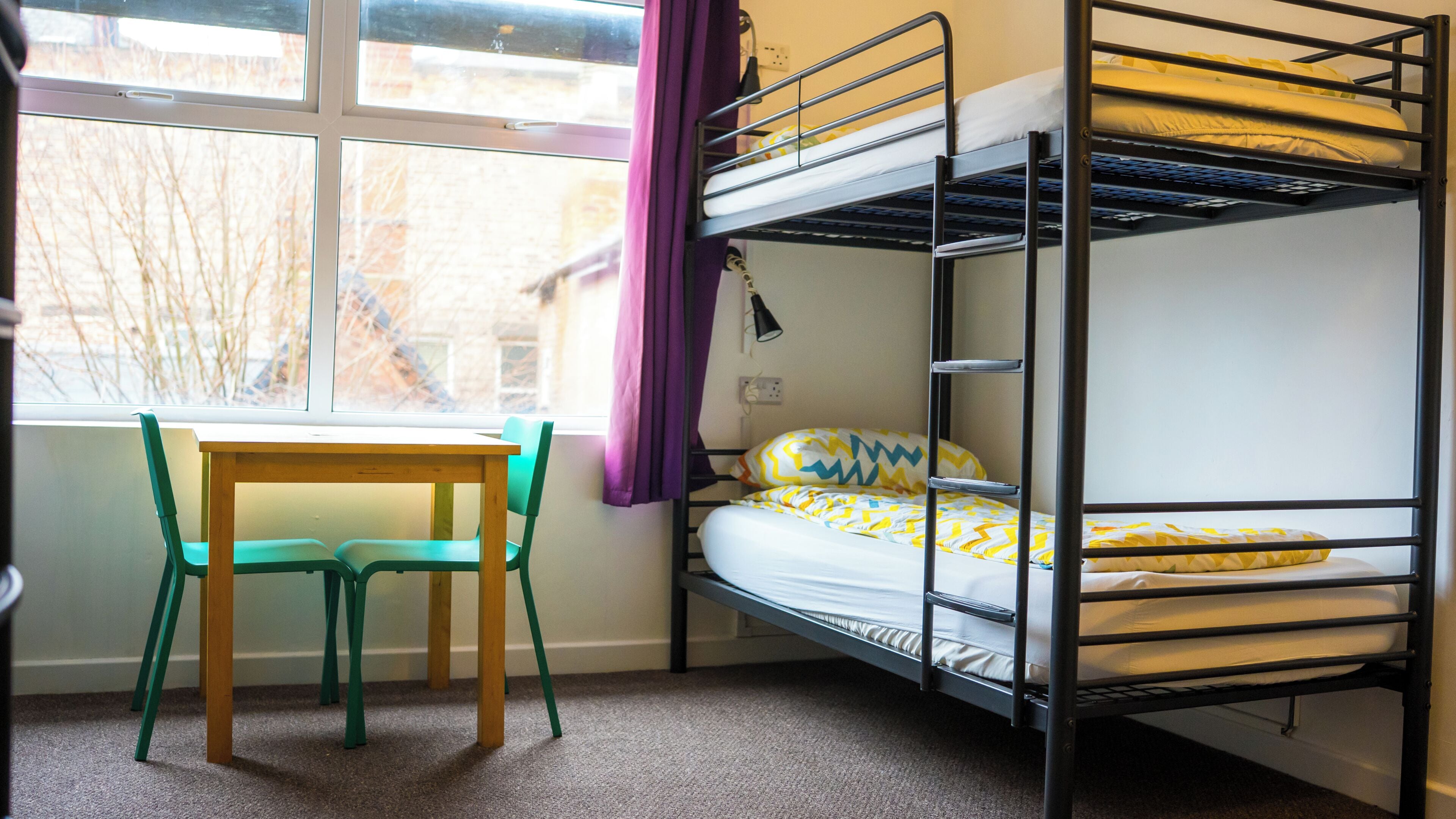 A bunk bed with a yellow and white patterned bedspread, a wooden table with two chairs, and a window with a view of bare trees.