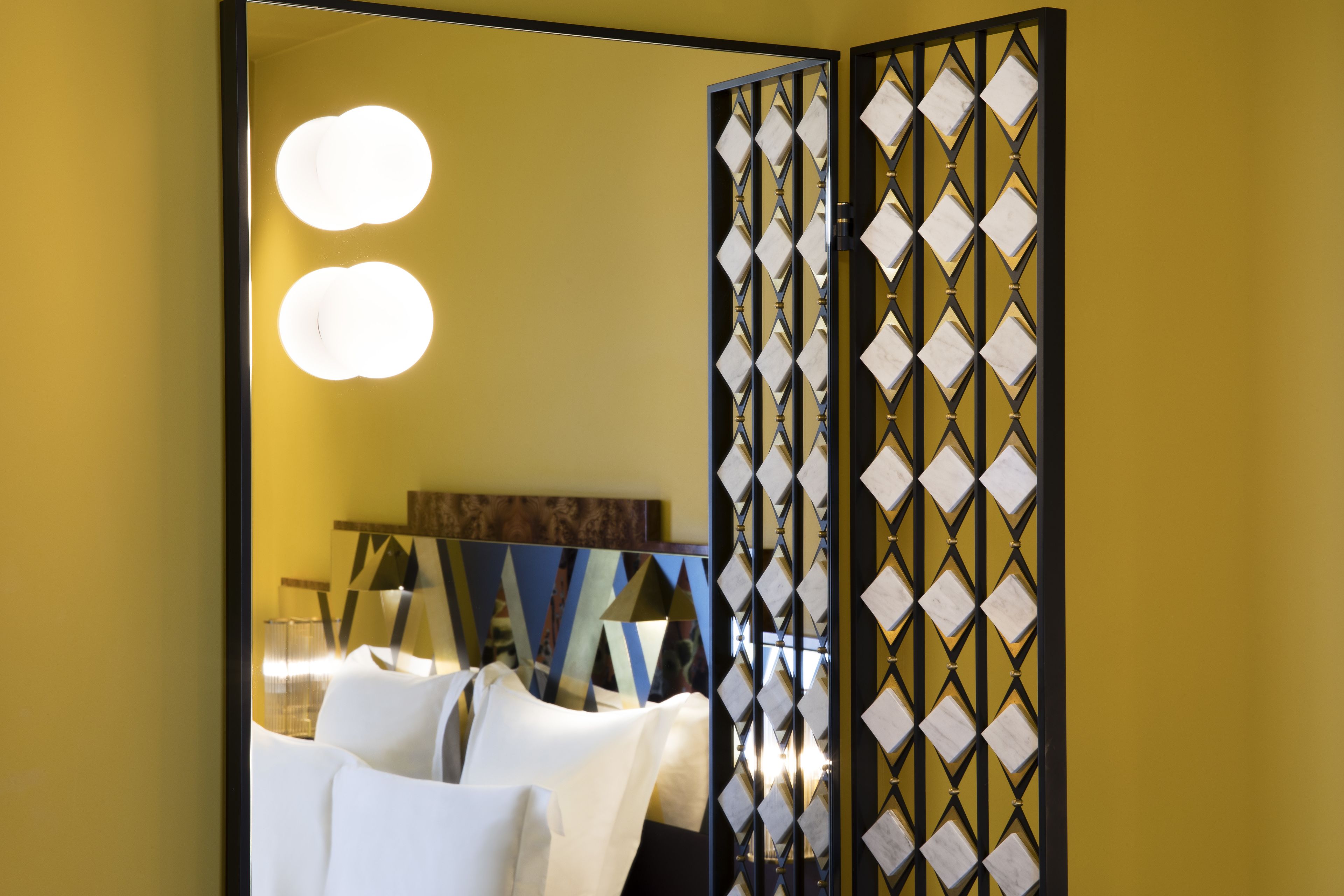 Habitación de hotel con un espejo grande, una cama con ropa de cama blanca y un separador decorativo con un diseño geométrico.