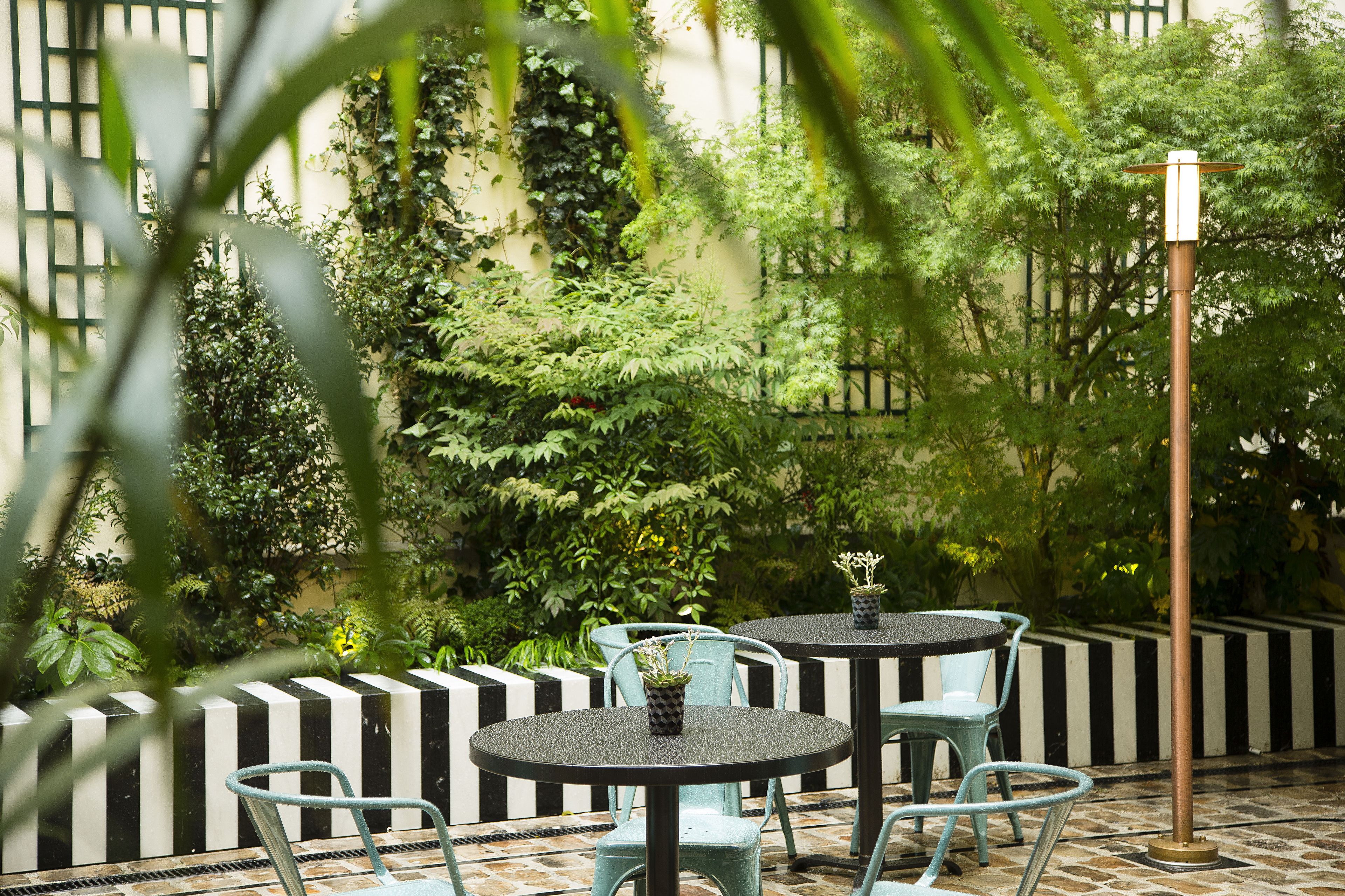 A patio area with round tables and chairs, surrounded by lush greenery and a striped fence.