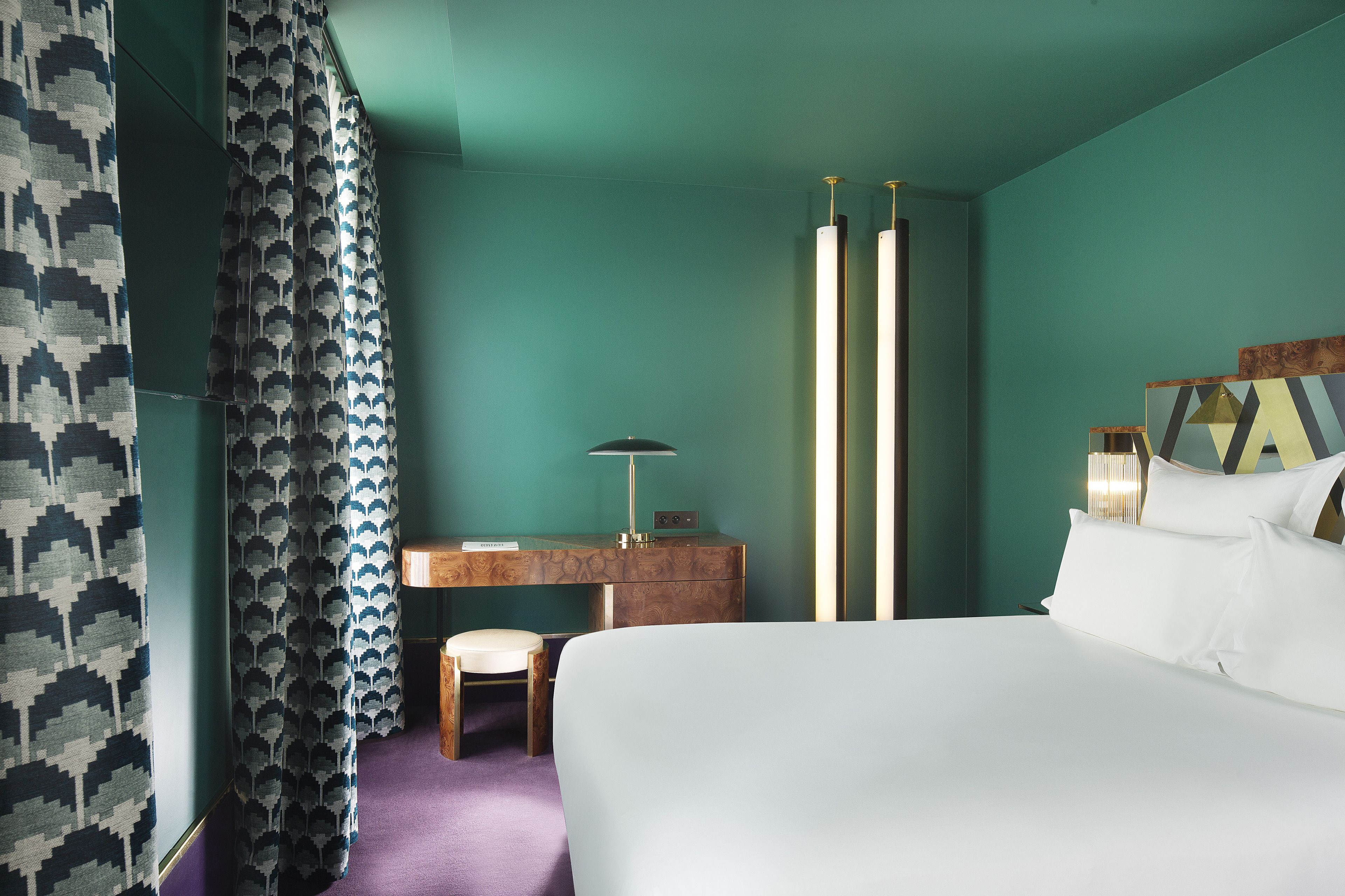 A hotel room with teal walls, a bed, a desk with a lamp, and a chair.