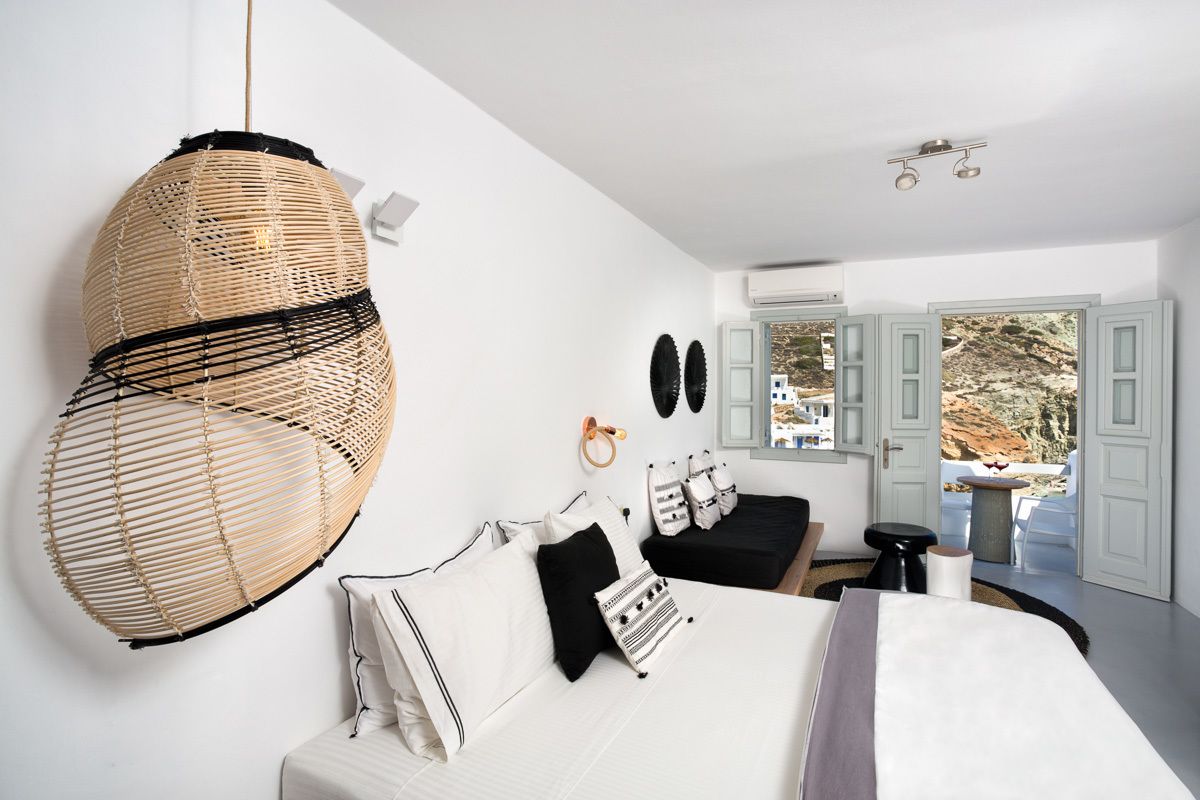 A modern bedroom with a large bed, a hanging rattan chair, a small table, and a view of the outdoors through open doors.