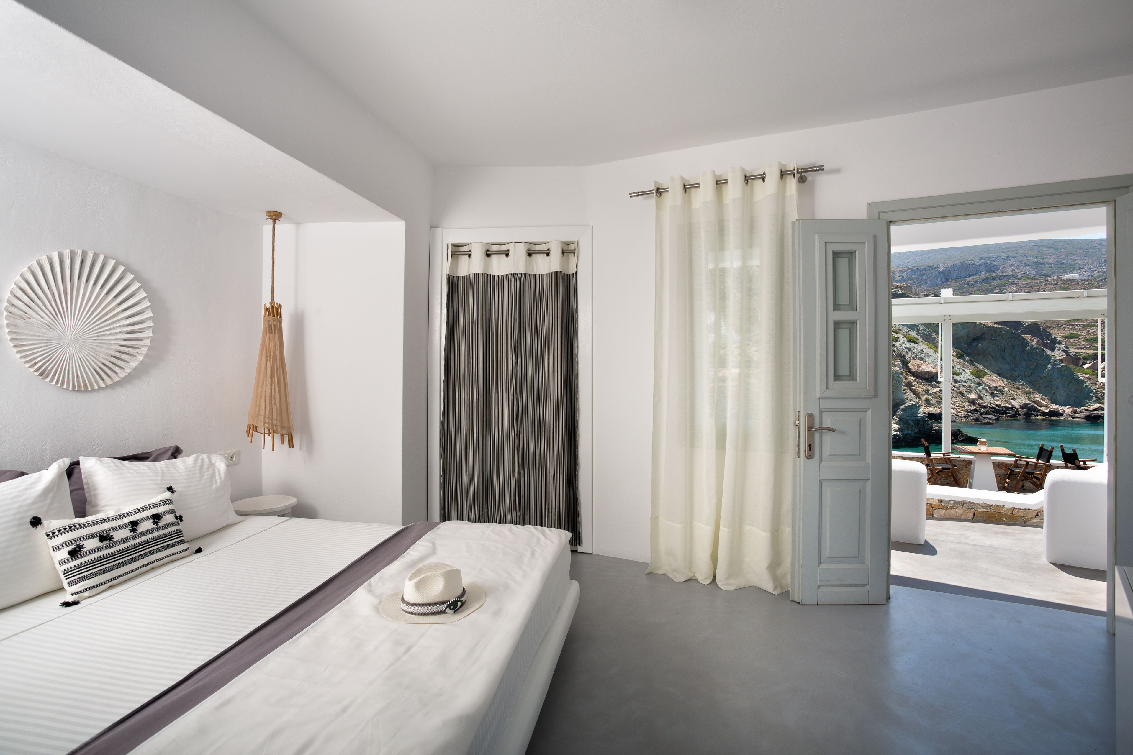 A bedroom with a bed, a view of a rocky coastline through an open door, and a balcony with outdoor seating.
