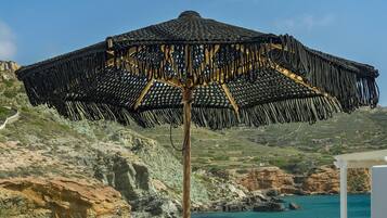 Un espacio de relax en la playa con una sombrilla tejida, dos sillones acolchados y una pequeña mesa con una bebida.