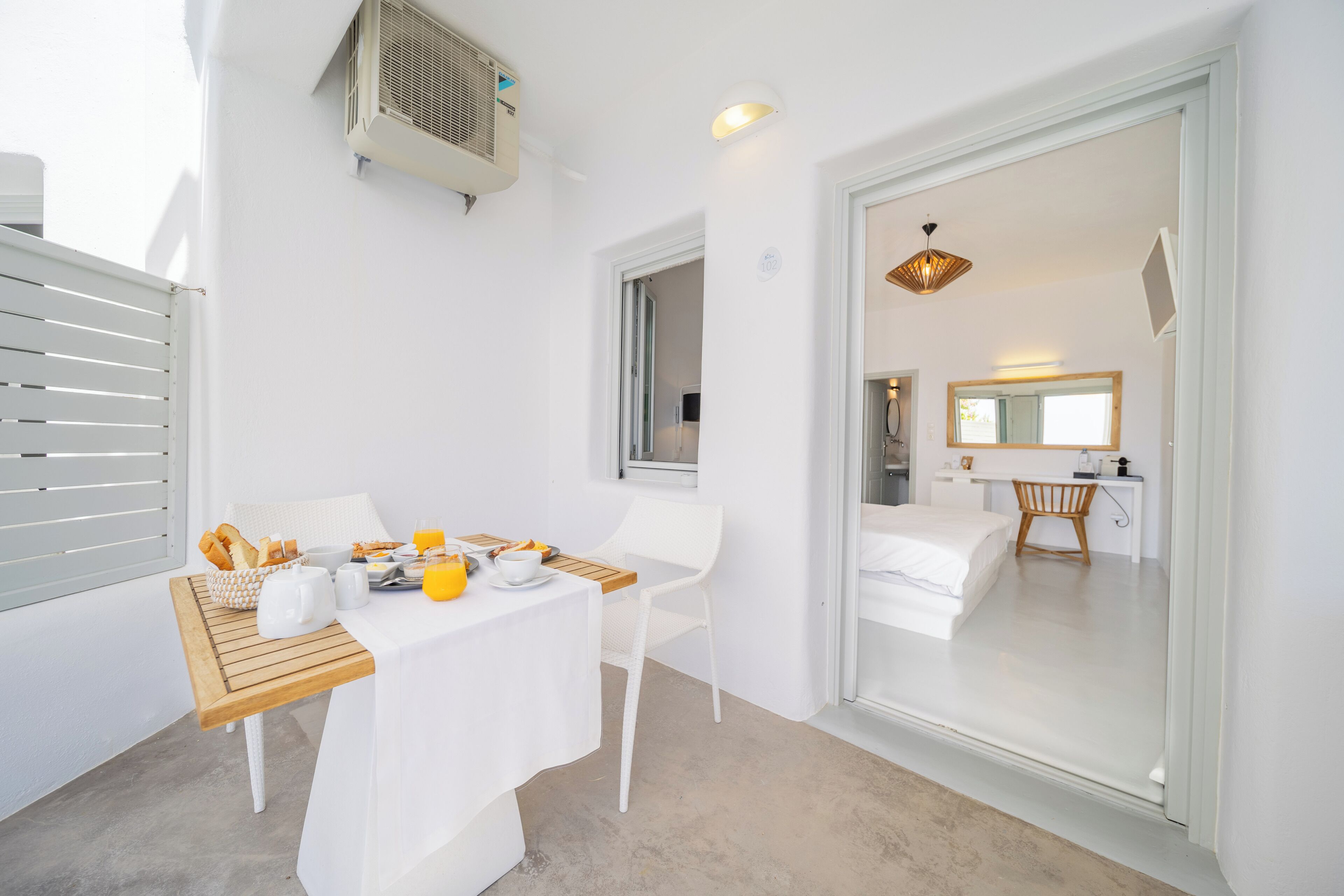 A modern room with a white table set for breakfast, a white chair, a bed with a wooden headboard, and a bathroom with a mirror and sink.