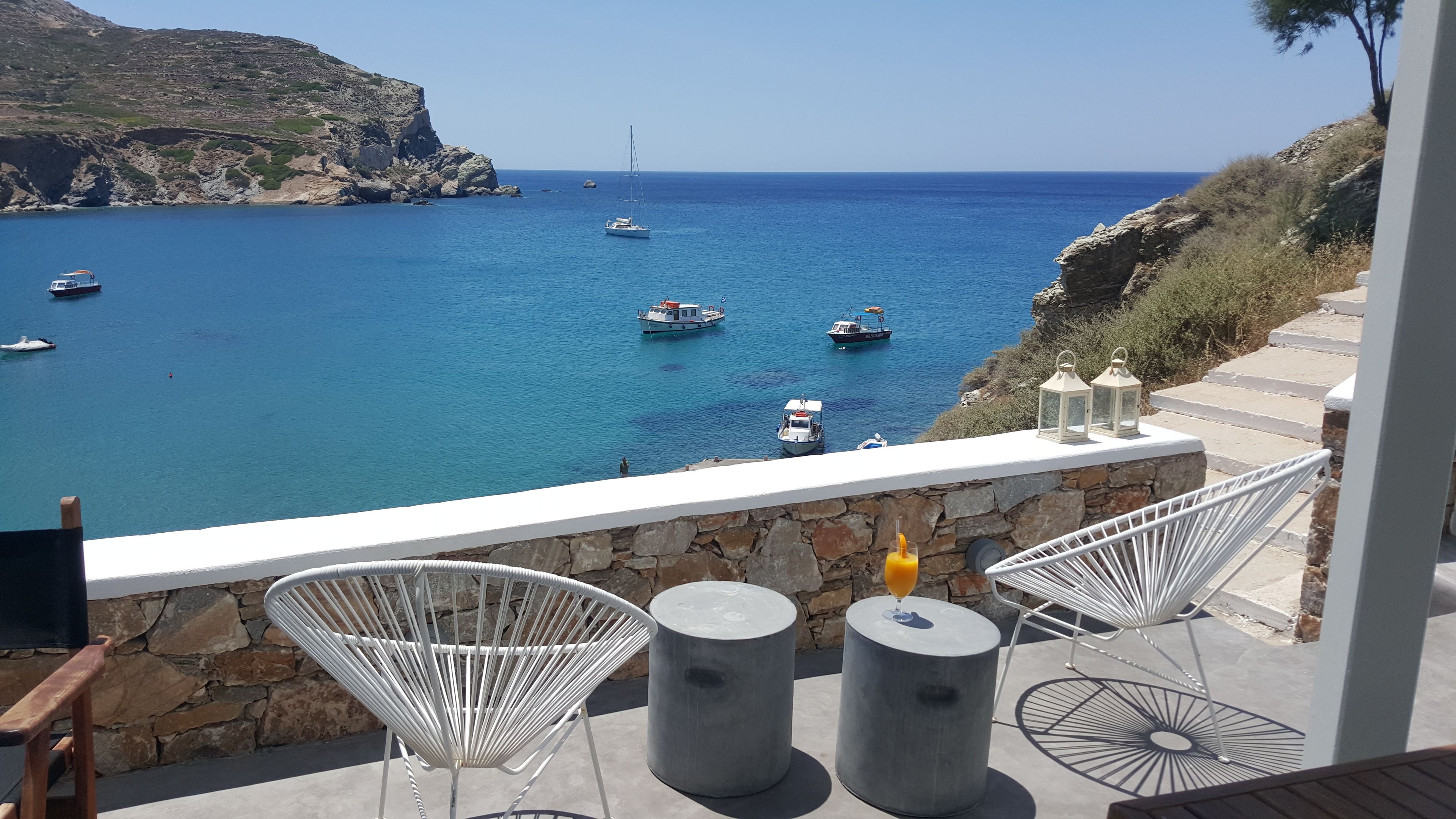 A terrace with white wicker chairs and two round tables, overlooking a coastal sea with boats.