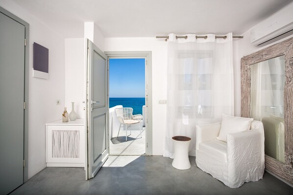 A modern room with a white chair, a small table, and a view of the sea through an open door.